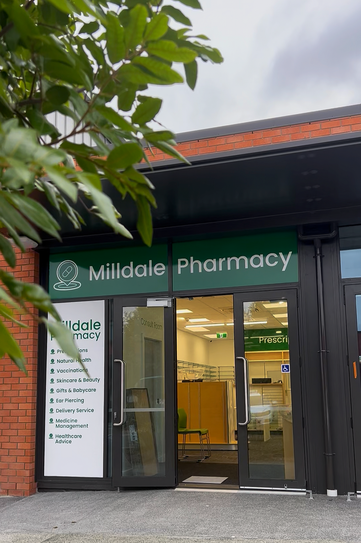Exterior of Milldale Pharmacy with glass entrance door, green signage, and a list of services on the side, surrounded by a red brick building.