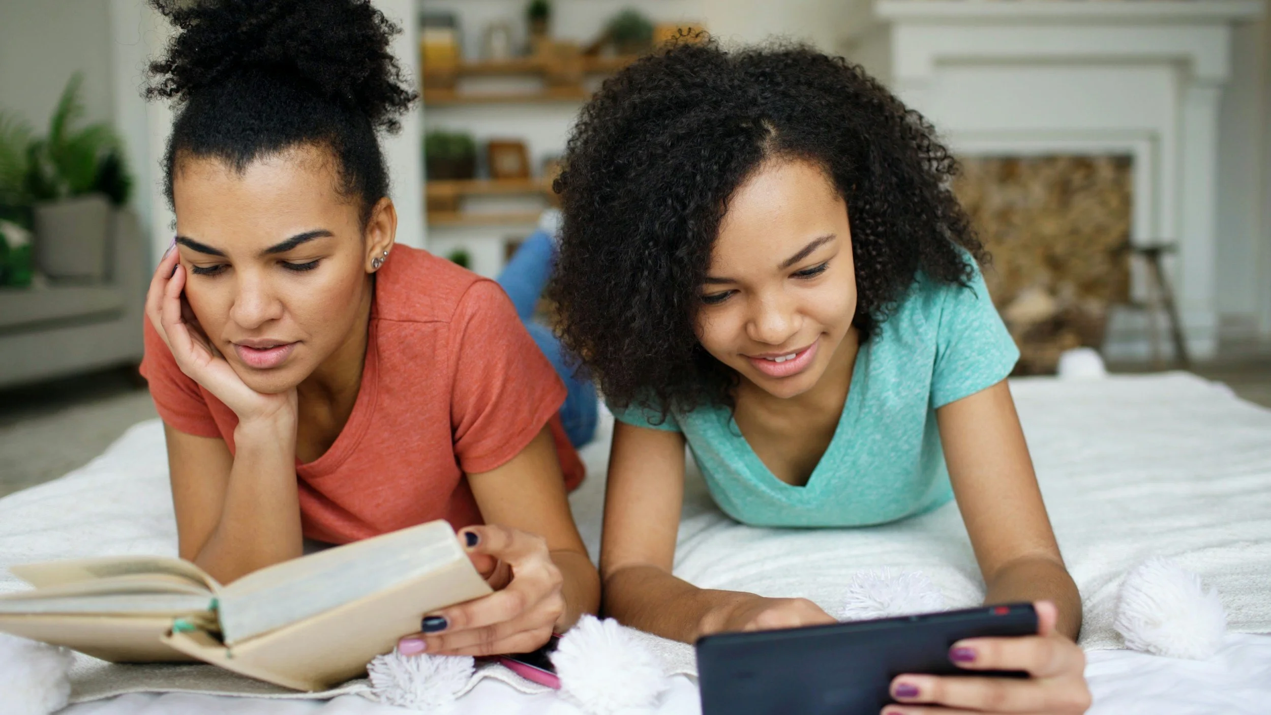 Two young women lying on their stomachs on a bed, looking at a tablet and a book, with a cozy living room in the background.
