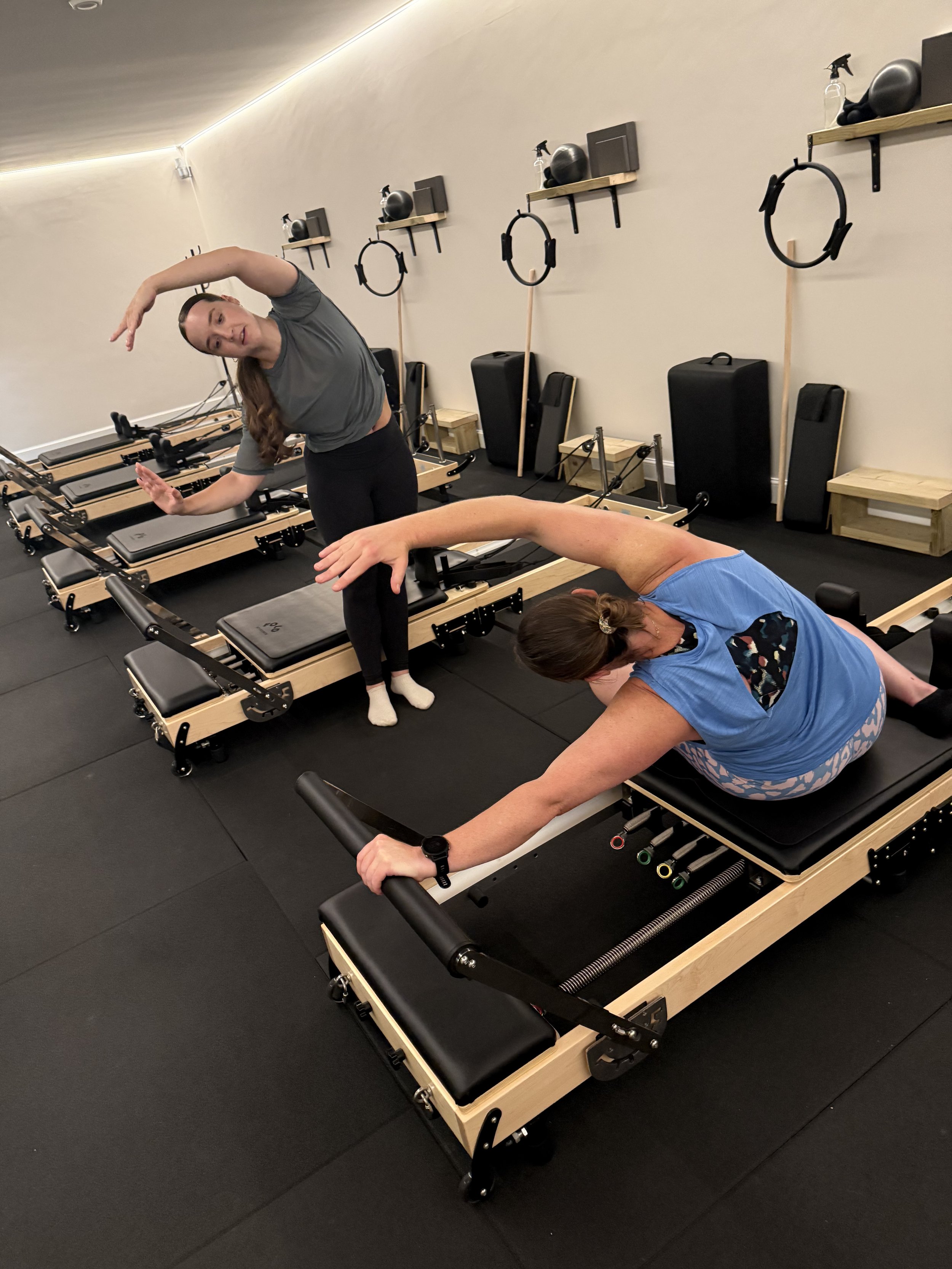 Two women practicing Pilates on reformer machines in a fitness studio. One woman stands next to a machine, demonstrating an exercise, while the other is lying face down on a machine, reaching forward with her arm.