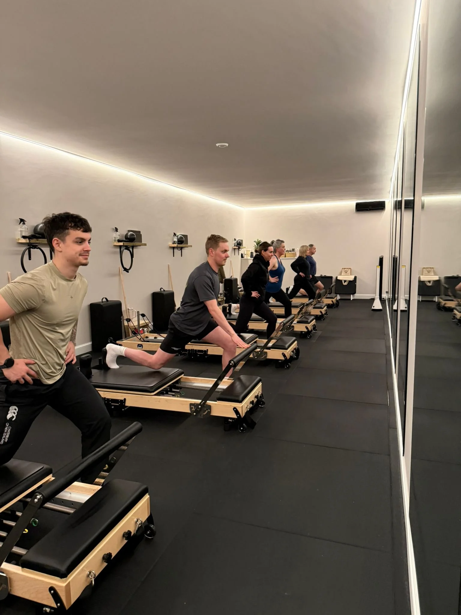 Group of people exercising on reformer Pilates machines in a fitness studio with mirrors and shelves on the wall.