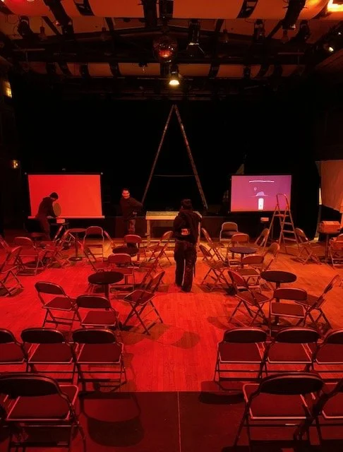 Empty theater space lit in deep red during the setup of Vibrations of Blackness, with scattered chairs, projection screens, and suspended sculptural elements forming a temporary ritual environment.