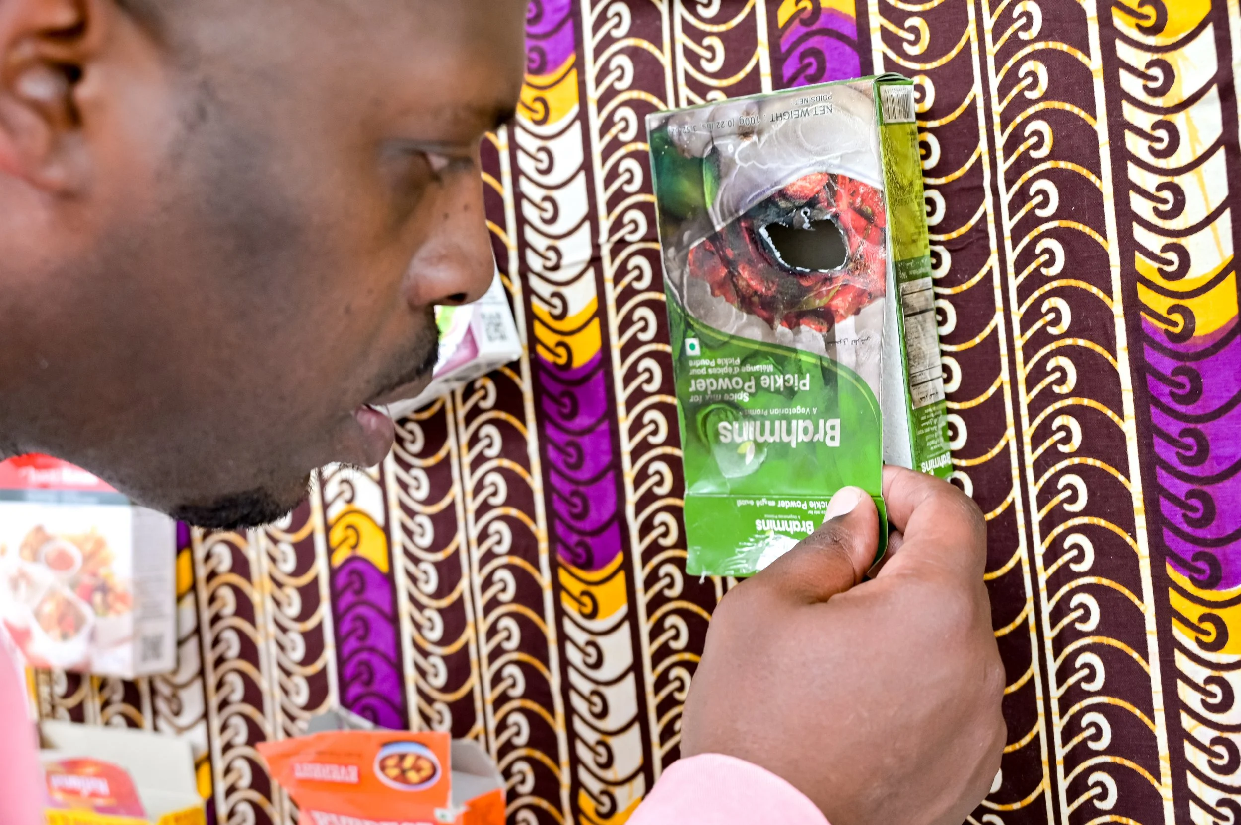 Close-up of Jonah Batambuze examining altered spice packaging mounted on patterned textile within an installation about diaspora, consumption, and ritual.