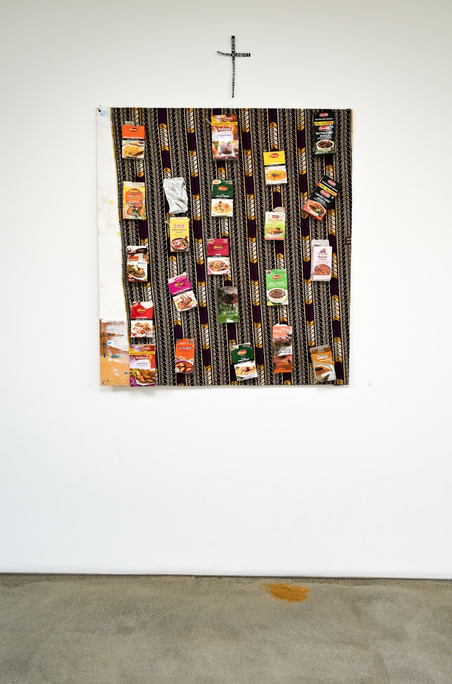 Mixed-media installation artwork featuring rows of South Asian spice packets mounted vertically onto patterned textile fabric beneath a small wall-mounted crucifix.