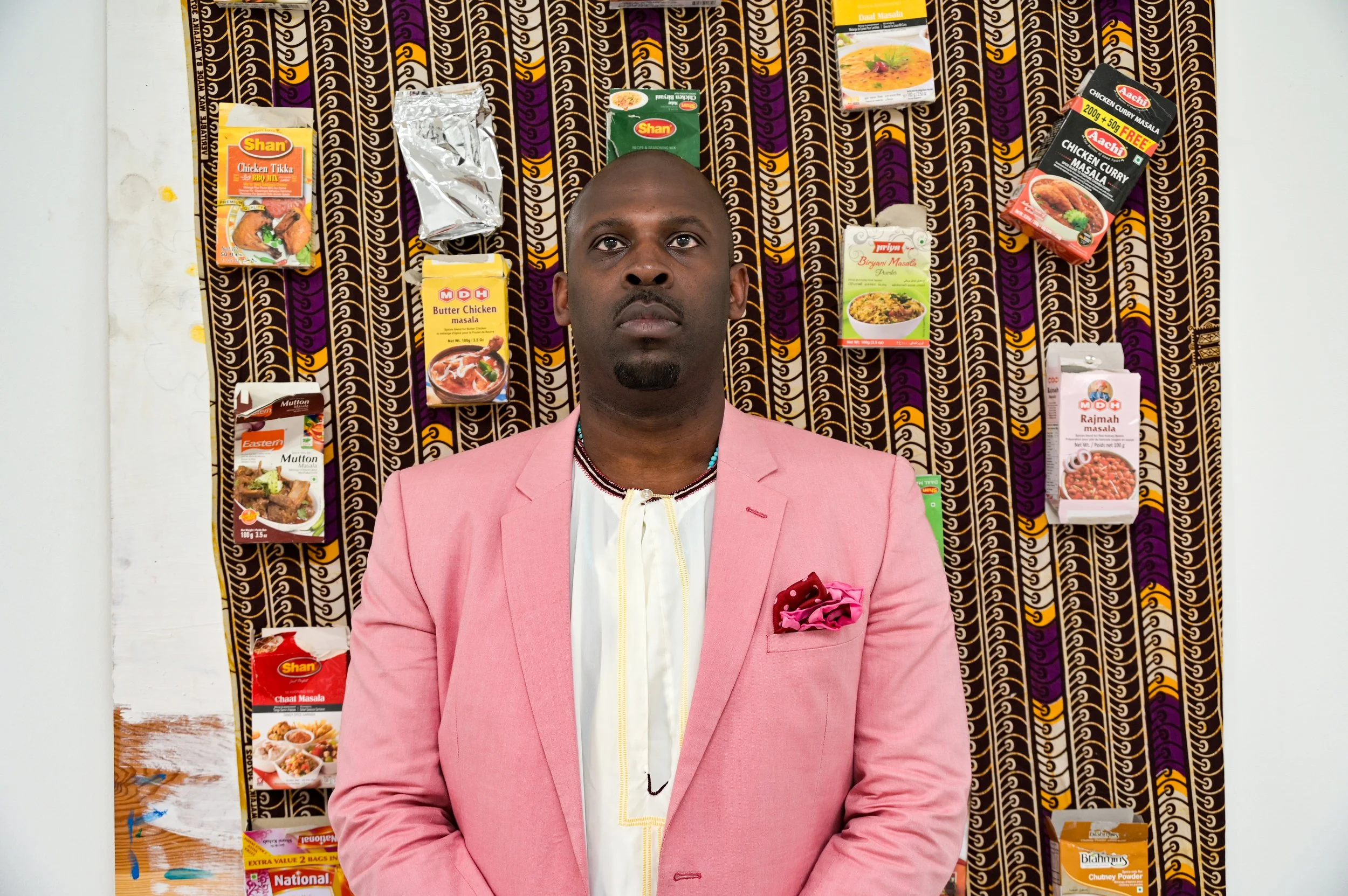 Portrait of Jonah Batambuze standing before an installation of South Asian spice packaging and patterned textile exploring migration, ritual, and diasporic identity.