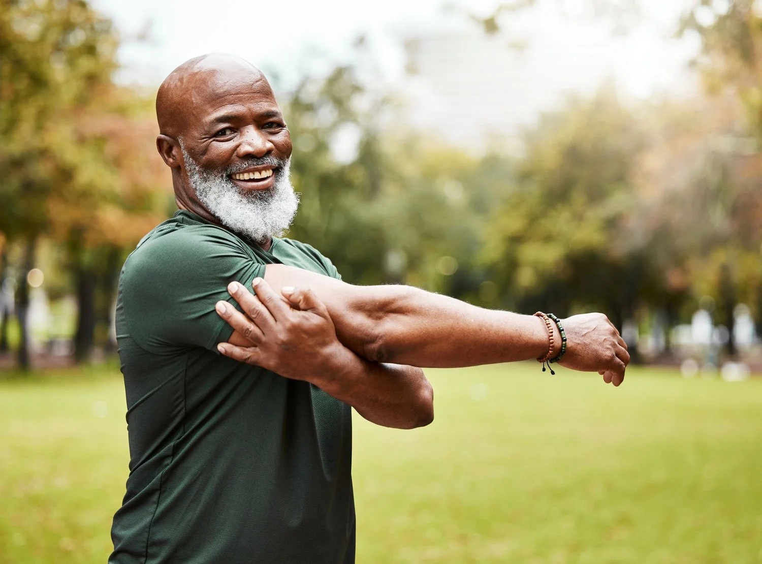 An elderly man with a gray beard wearing a green sports shirt stretching his arm in a park with trees and grass in the background. SNAPPS - Comprehensive General Practice Healthcare Articles.