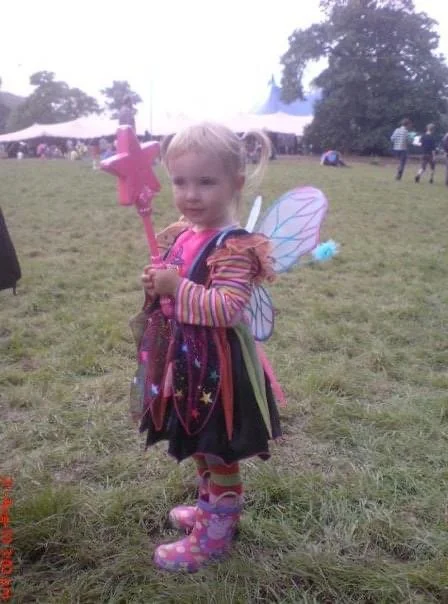 Young girl dressed as a fairy with butterfly wings, holding a pink star-shaped wand, standing outdoors on grass during a festival or event.