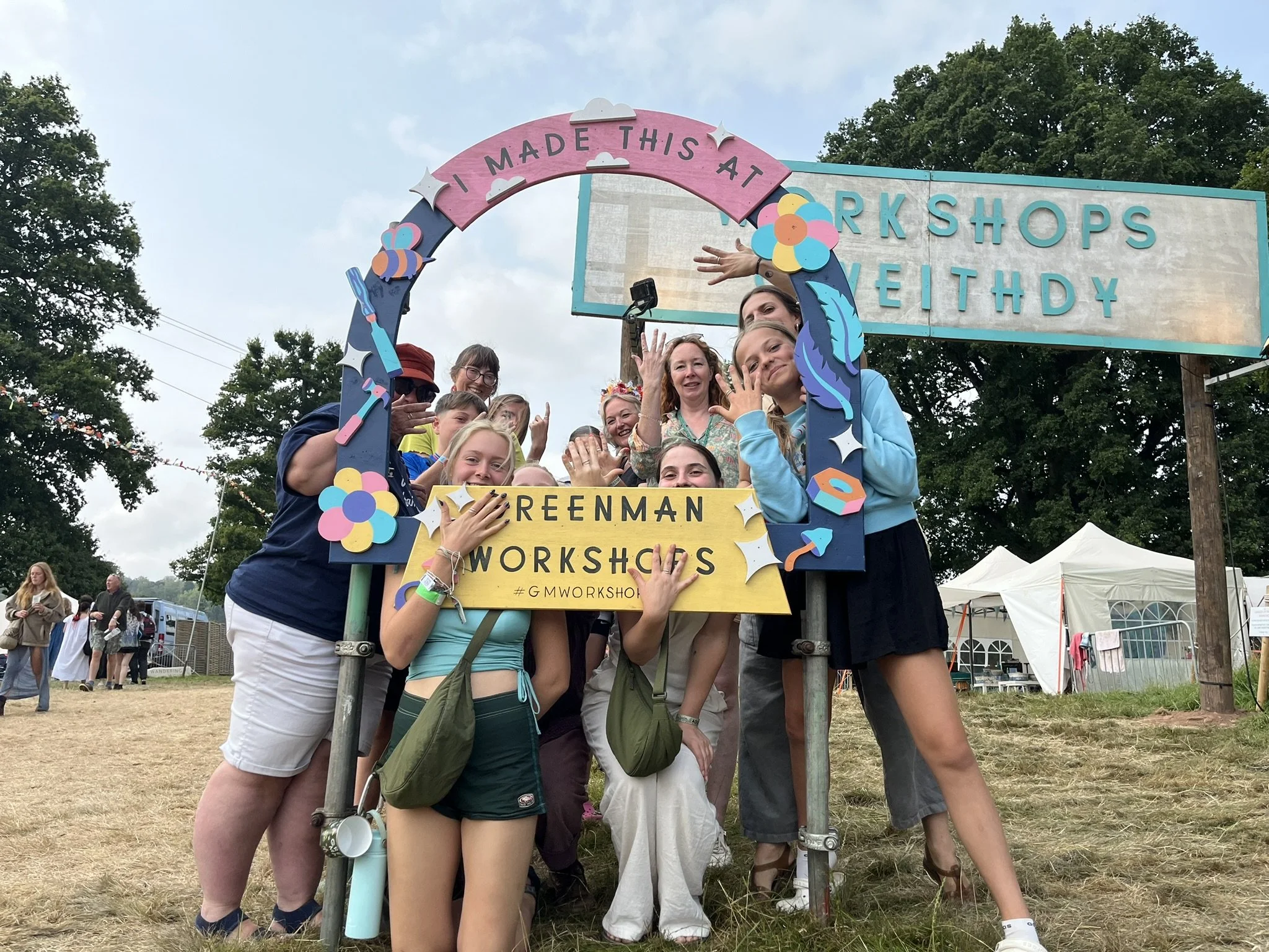 Group of people at the Greenman Workshops at a festival, standing behind a decorative frame with colorful flowers and craft supplies, under a sign that says 'Greenman Workshops' and 'I Made This At Workshops Weithdy'.