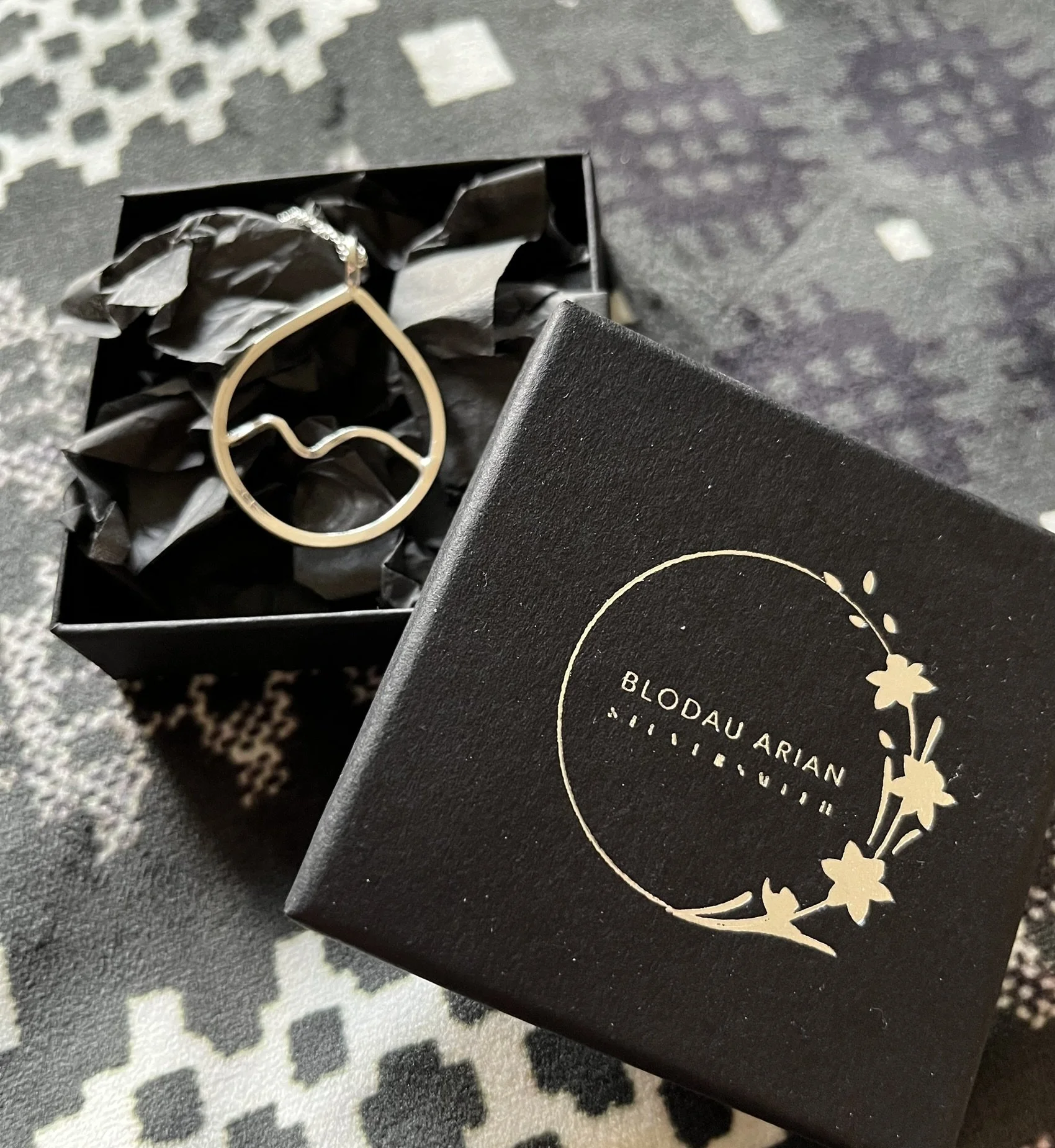 A jewelry box containing a silver necklace with abstract wave design, next to a black box with gold floral decoration and the name Blodau Arian, placed on a patterned surface.