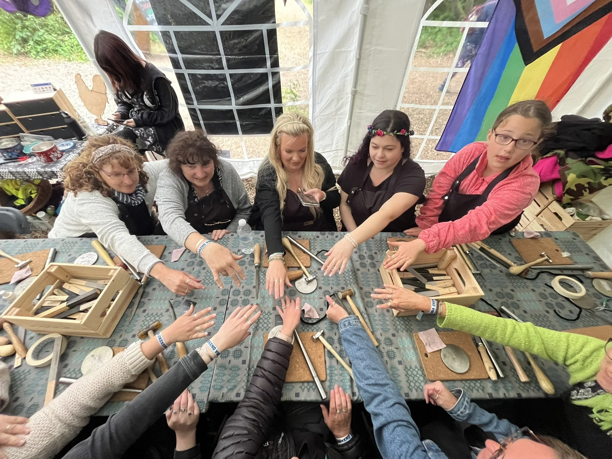 Group of women at a craft workshop, reaching towards the center of a long table with woodworking tools, wooden boxes, and crafting supplies inside a tent with a rainbow flag in the background.