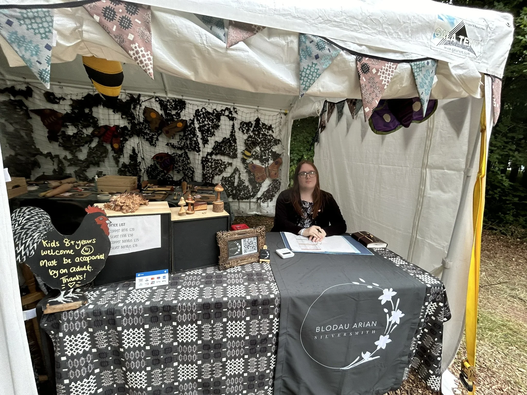 A woman sitting at a booth under a white canopy tent decorated with bunting, with butterfly and insect craft displays behind her, for a craft or market event.