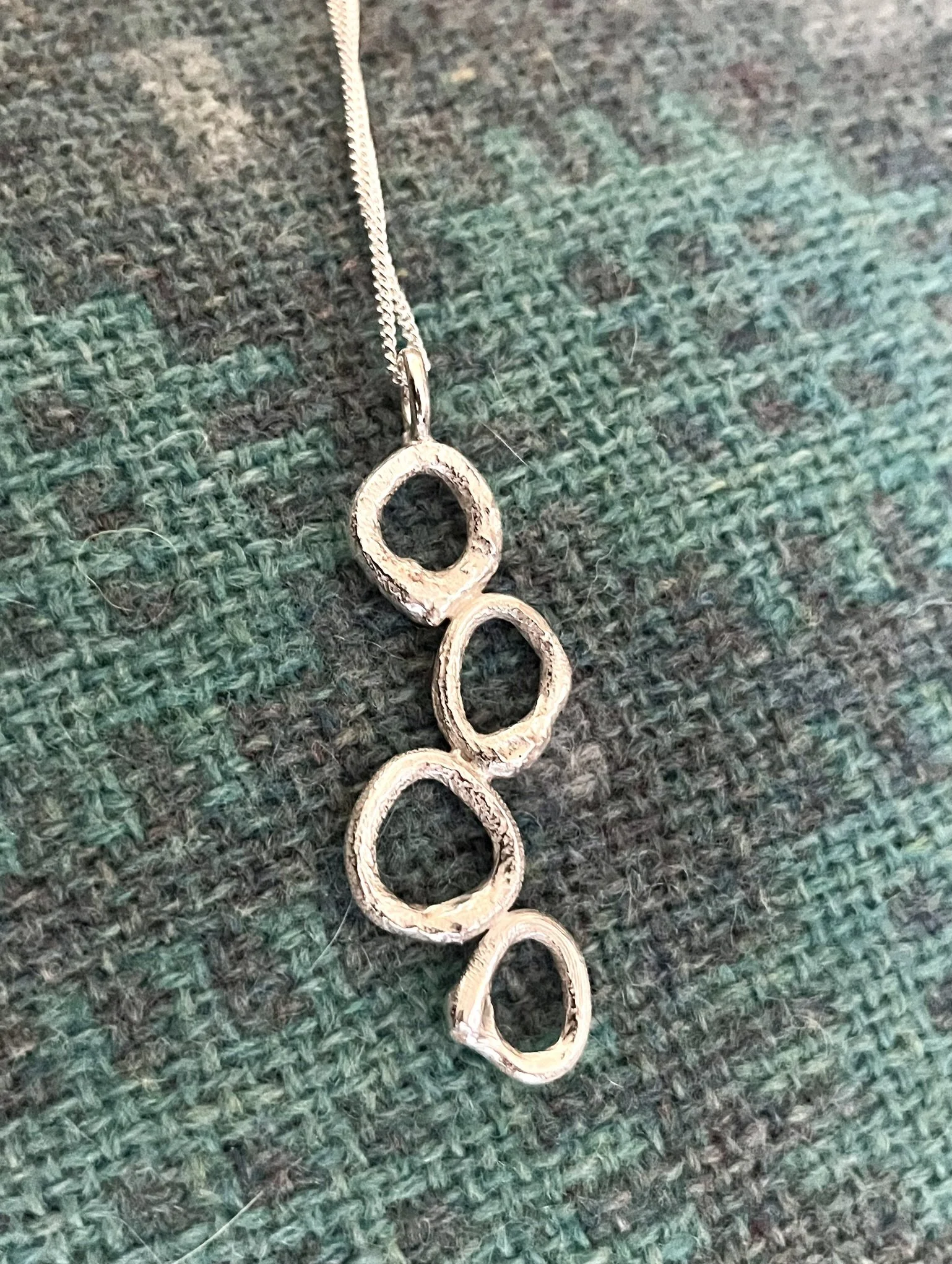 Silver necklace with four interconnected organic oval-shaped open loops, lying on a Welsh tapestry blanket.