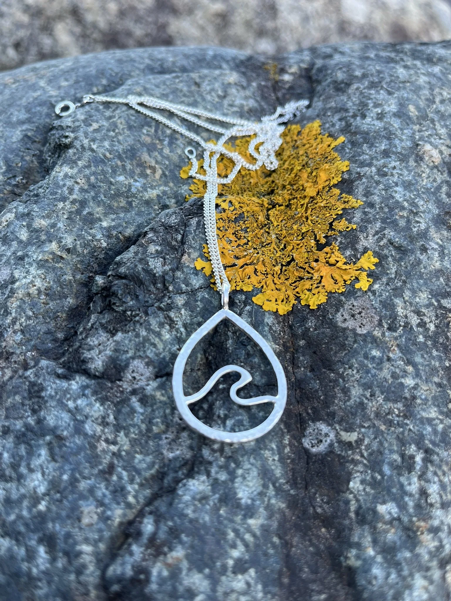 A silver necklace with a pendant featuring a wave design, resting on a gray rock with yellow lichen.