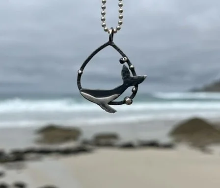 Necklace with a fish-shaped pendant hanging on a chain, beach and ocean in the background under cloudy sky.