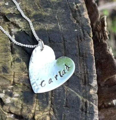 A silver heart-shaped pendant with the name 'Carla' engraved on it, hanging on a silver chain against a wooden background.