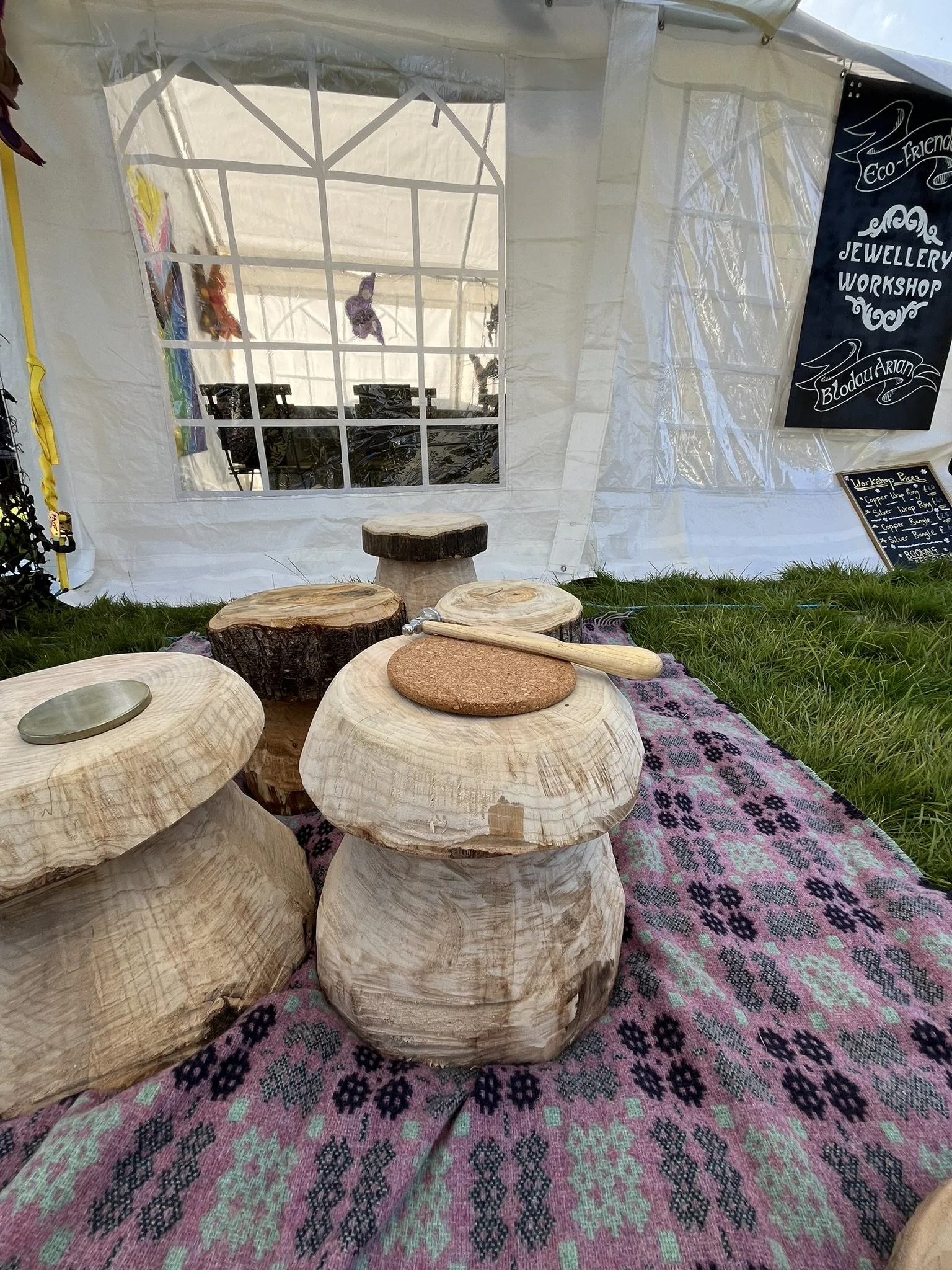 Wooden craft stools arranged on patterned fabric at an outdoor wedding fair booth, with a mirror, a welcome sign, and a blackboard in the background.