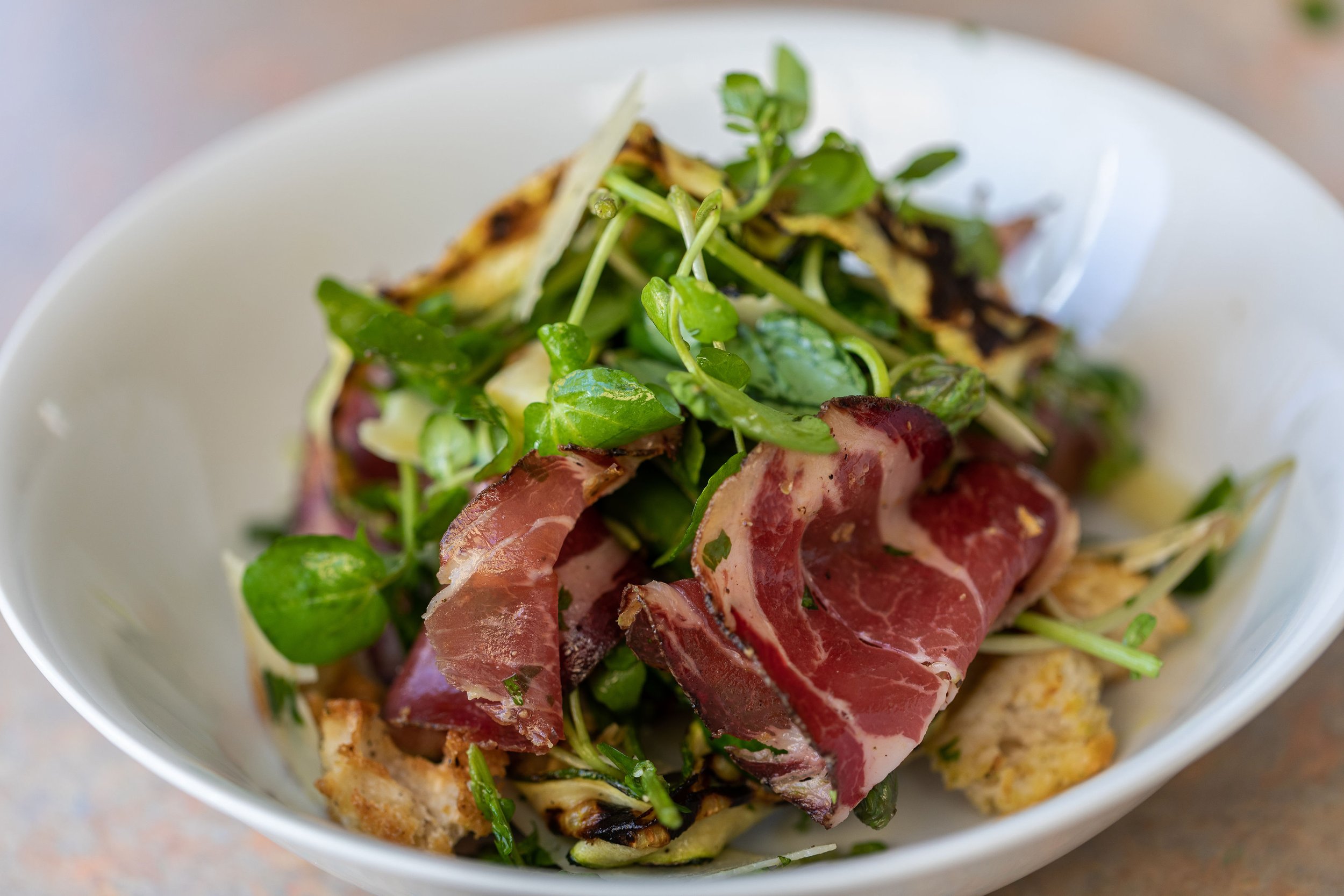 Salad with microgreens, grilled vegetables, and slices of prosciutto in a white bowl.