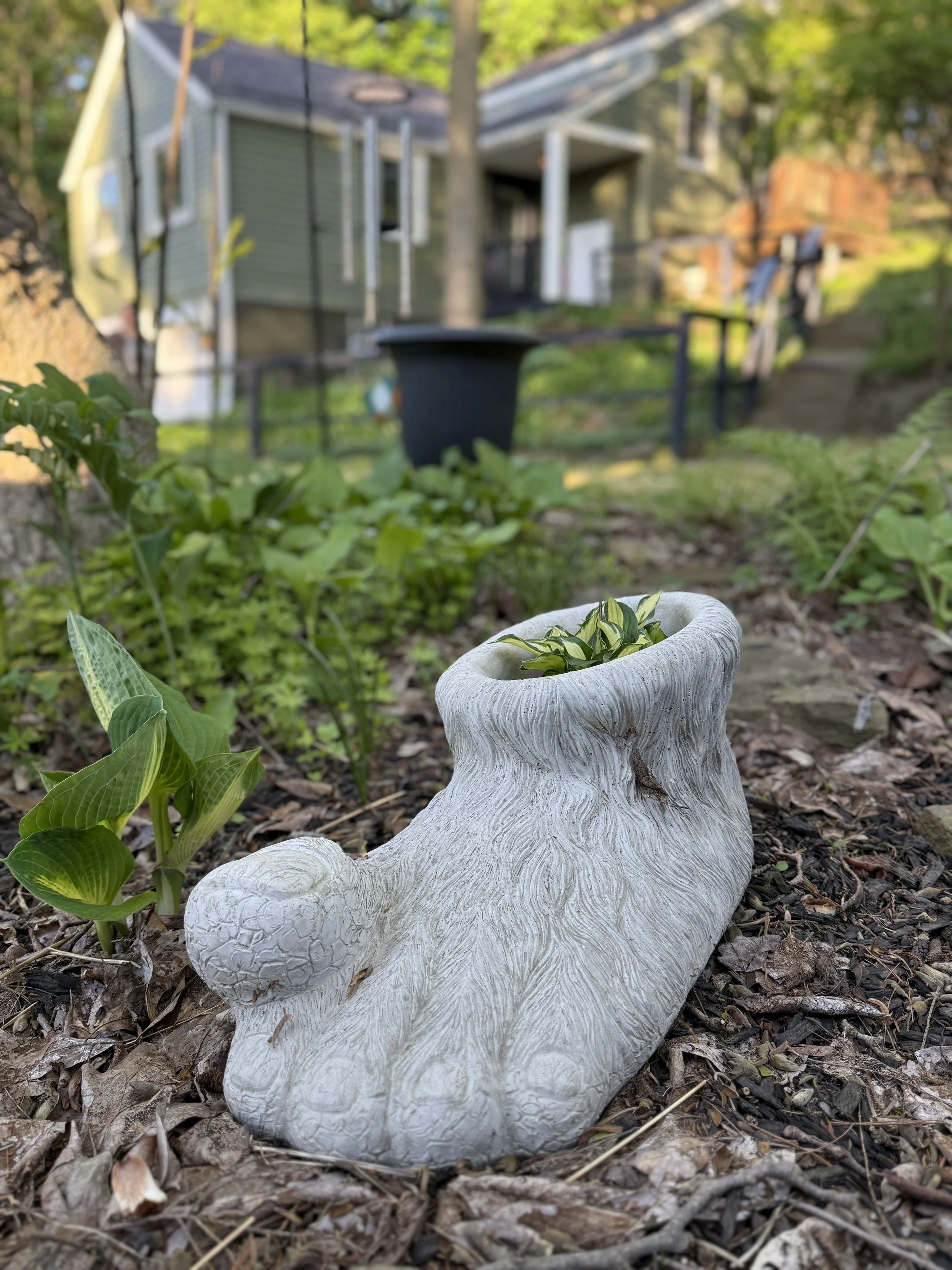 A decorative white stone hare-shaped planter lying on its side in a garden, with green plants nearby and a house with a porch and trees in the background.