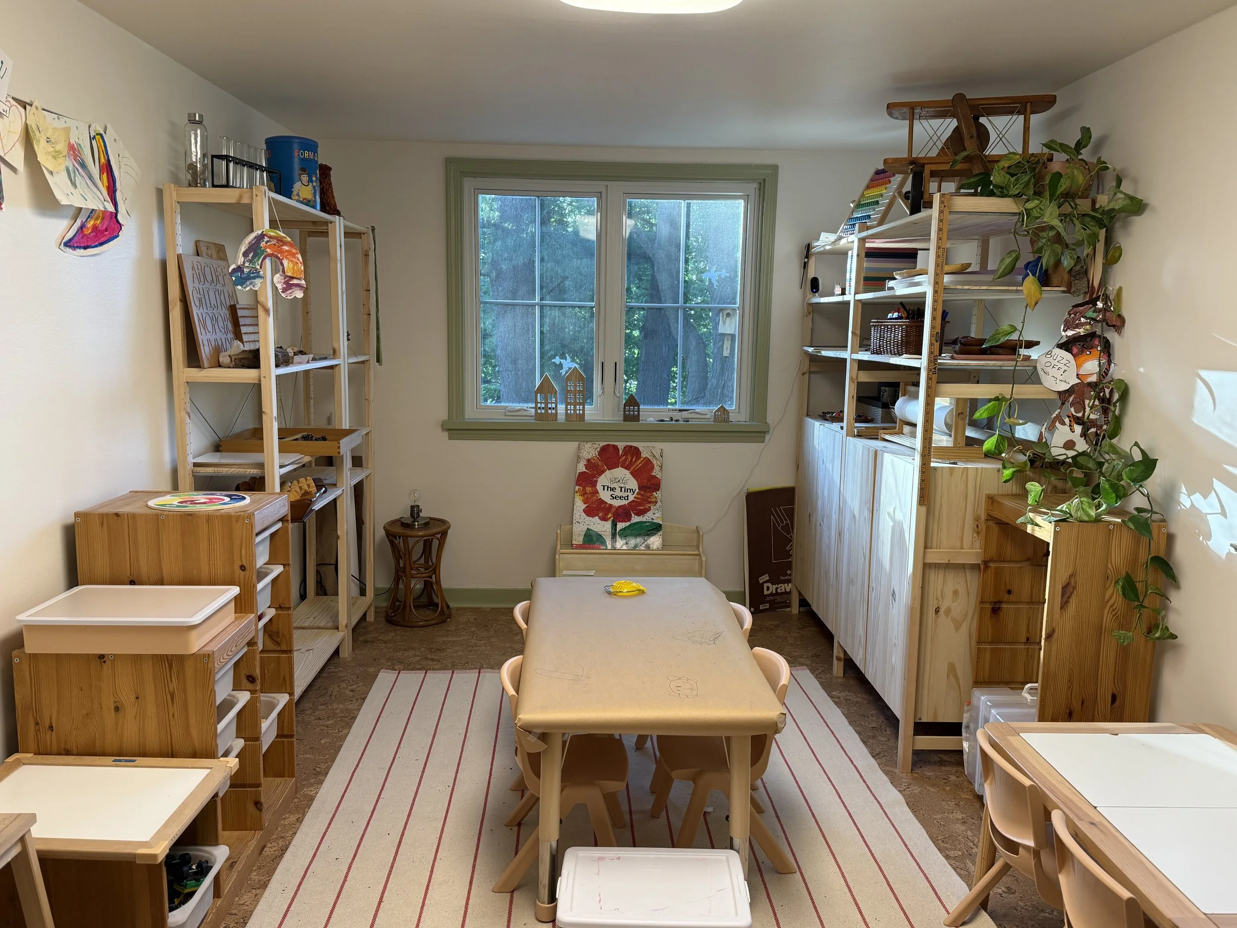 A small art or craft room with wooden shelves filled with art supplies, a window with greenery outside, and a table with chairs in the middle.