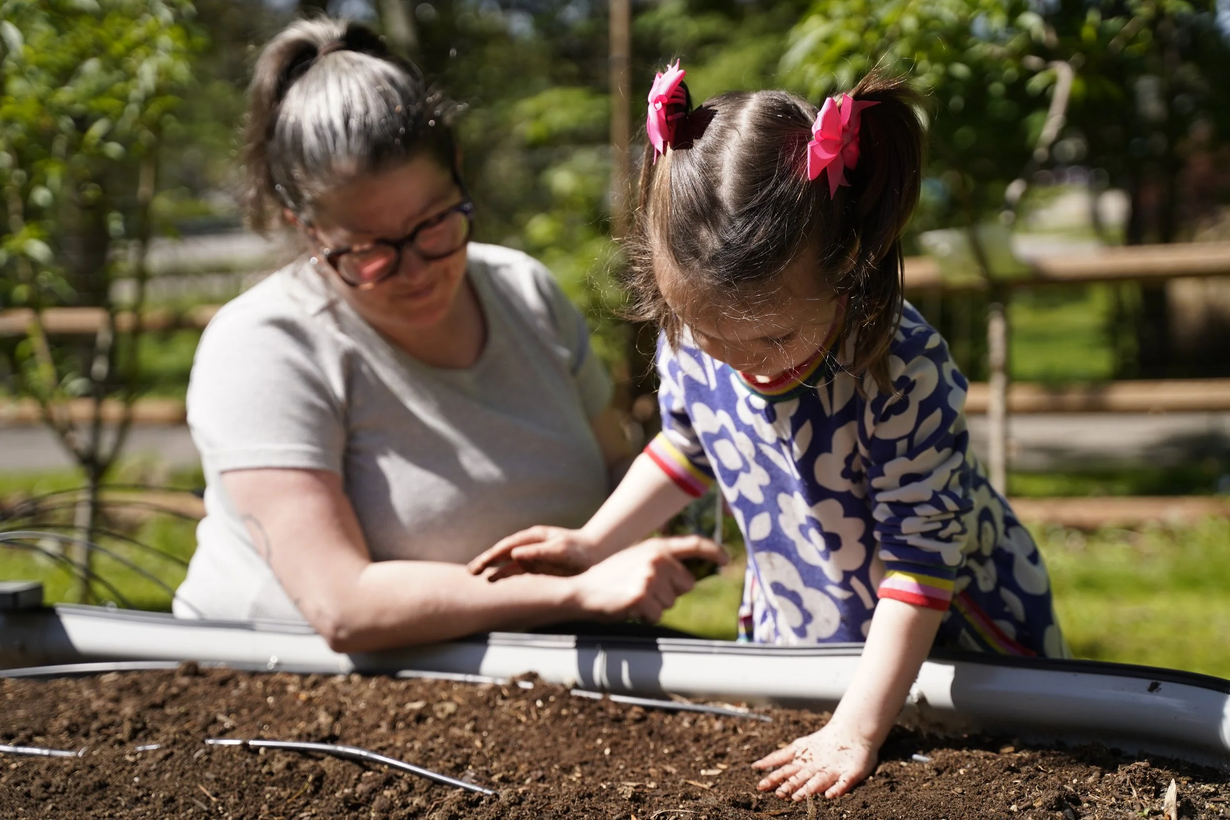 A woman and a young girl planting a seed in soil using a gardening tool outdoors on a sunny day.
