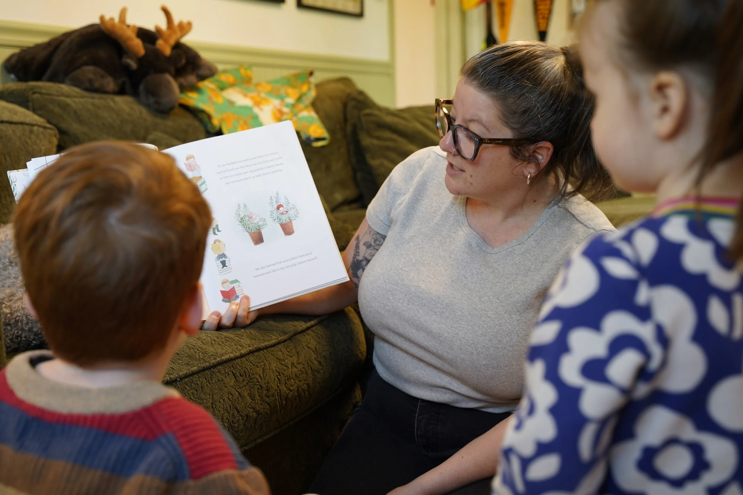 A group of children and a woman reading a colorful picture book together in a cozy room with a couch and decorative pillows.