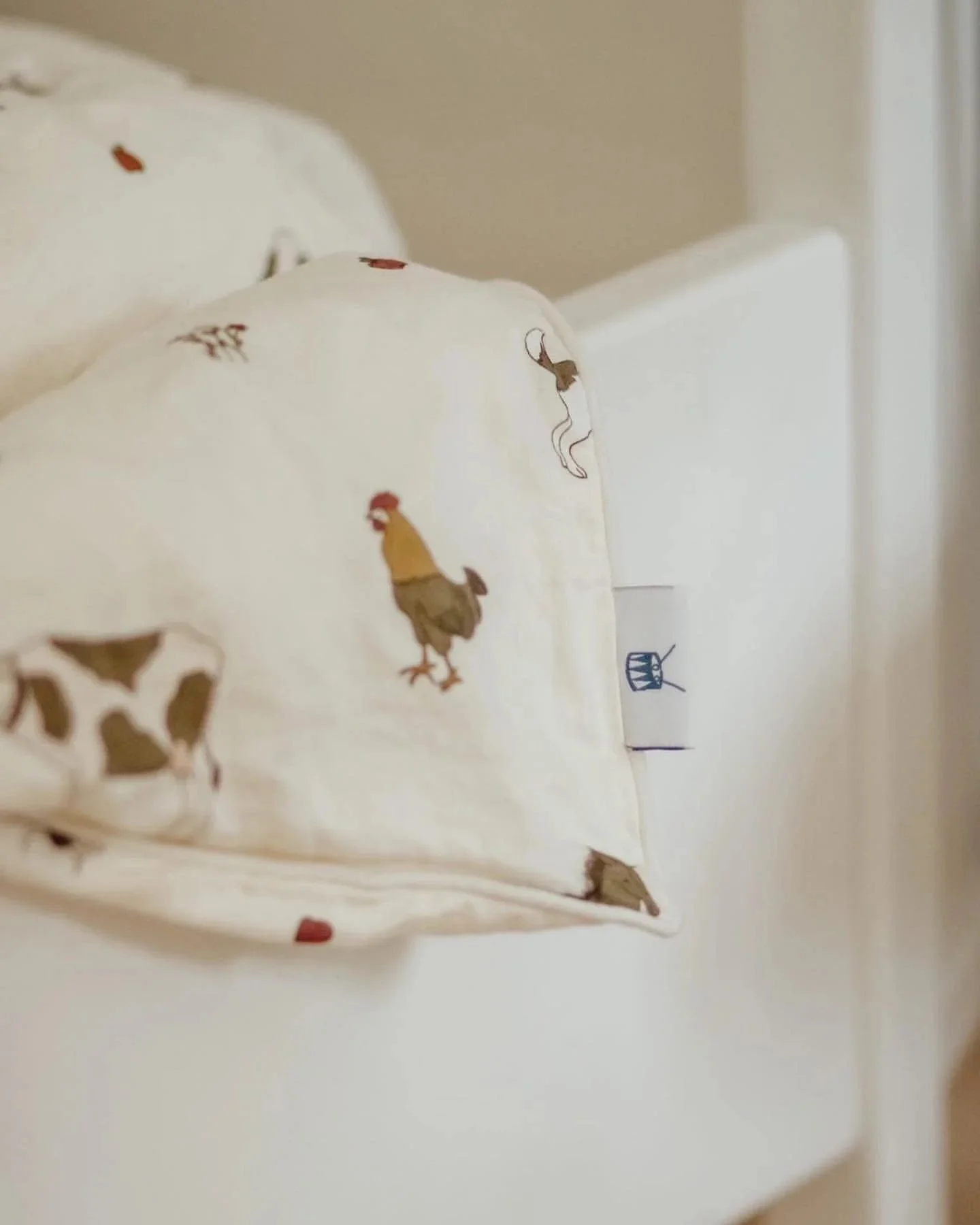 Close-up of a pillow with chicken and rooster embroidered patterns on white fabric, placed on a white bed with a beige wall in the background.