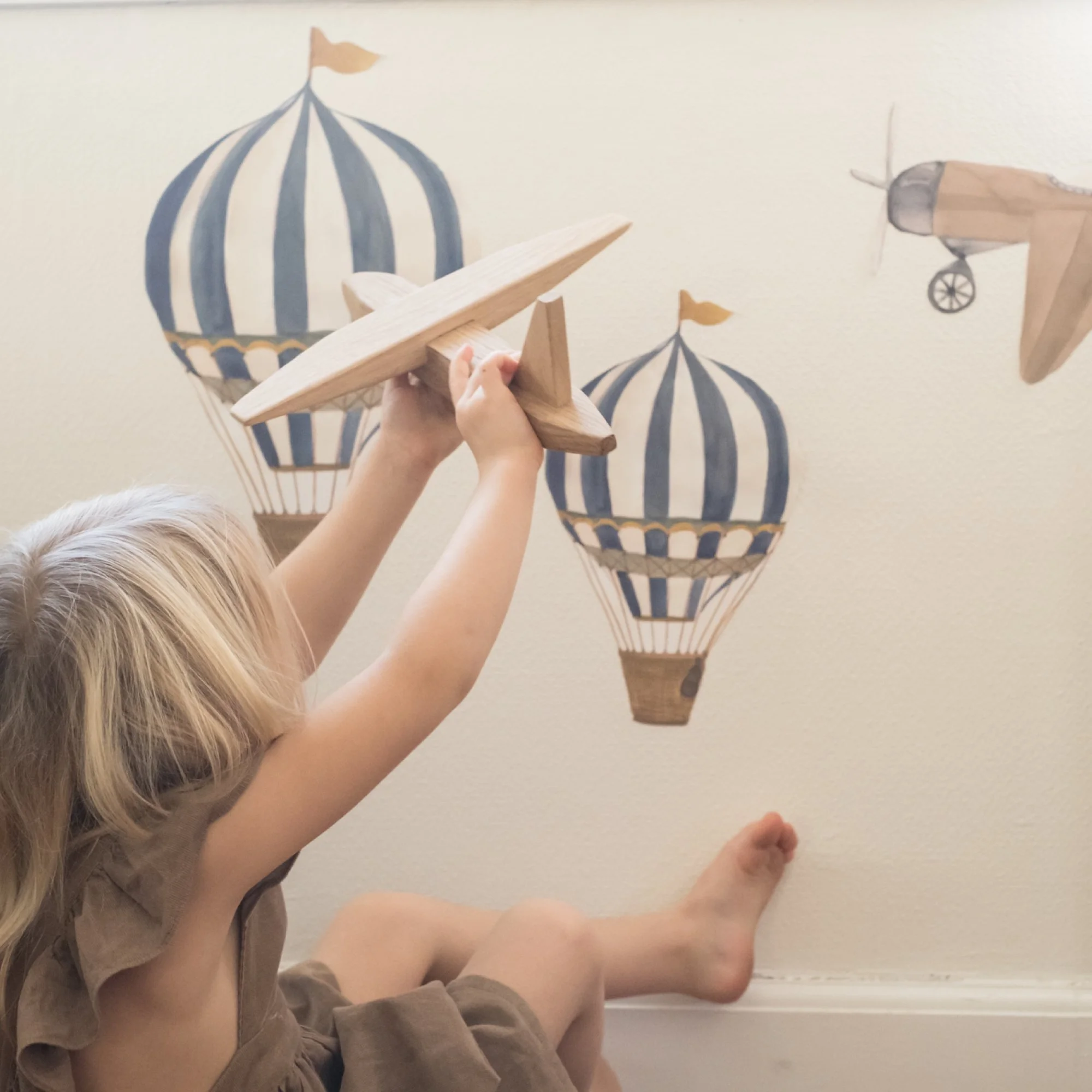 Child holding a wooden toy airplane while sitting on the floor in front of wall decorated with wall stickers with hot air balloon and airplane illustrations.
