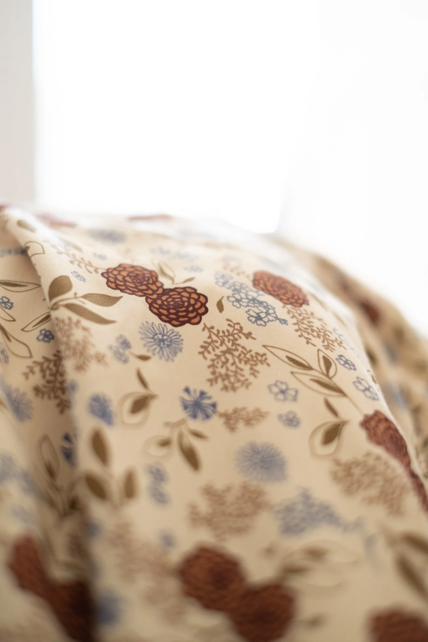 Close-up of a fabric with a floral pattern featuring bordeaux, light blue, and beige flowers and leaves on a cream background as bedding for that's mine