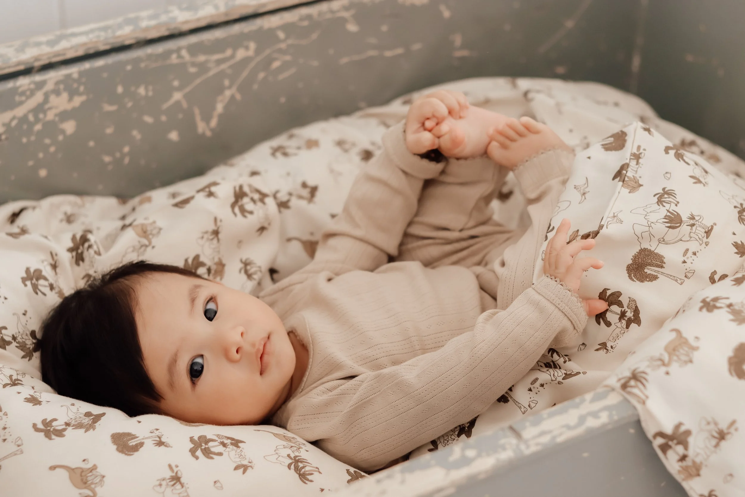 A young child with dark hair lying in a crib on a patterned blanket, looking at the camera with a neutral expression.
