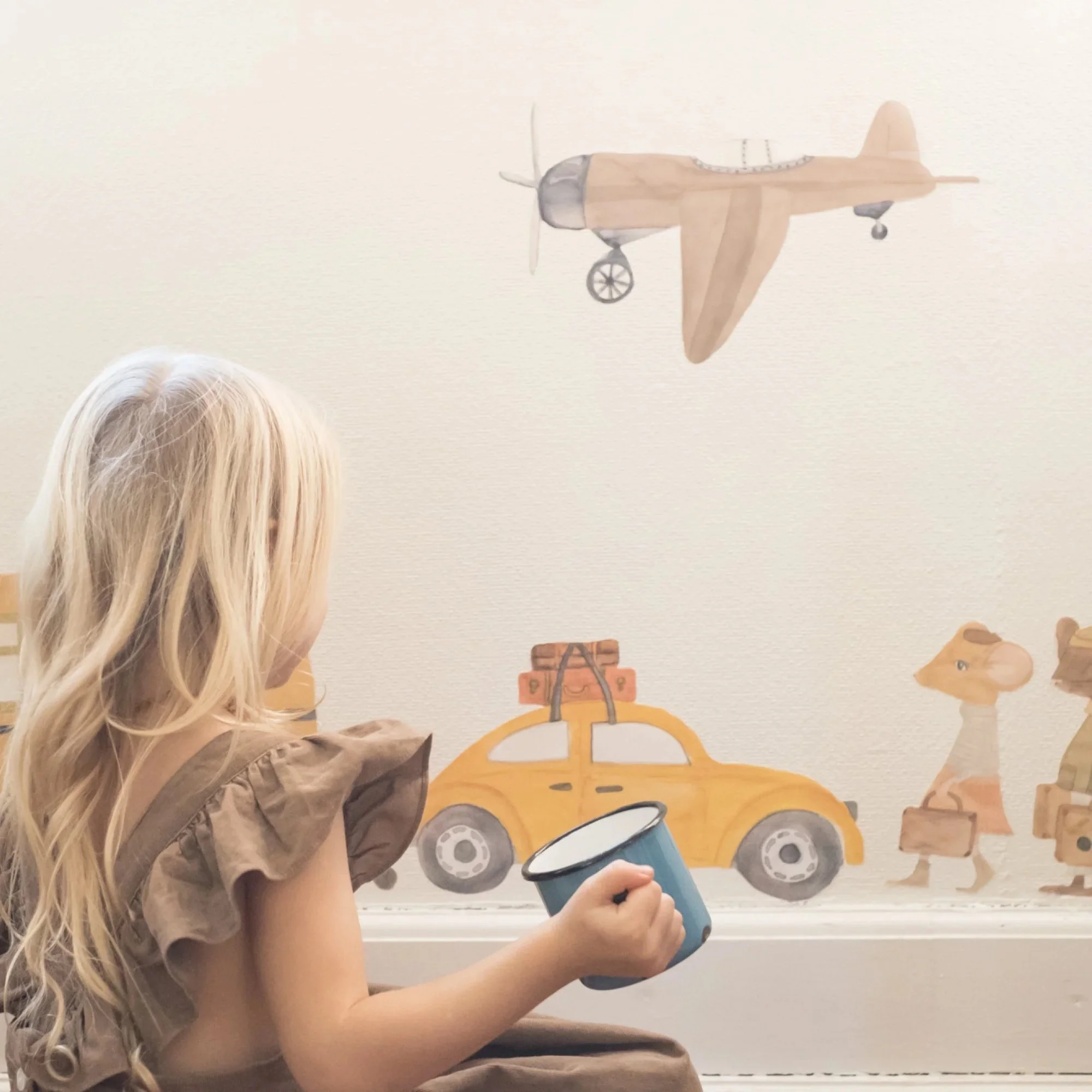 A young girl with long blonde hair sitting on the floor, holding a mug, facing a wall decorated with wall stickers airplane, car, and animal illustrations.