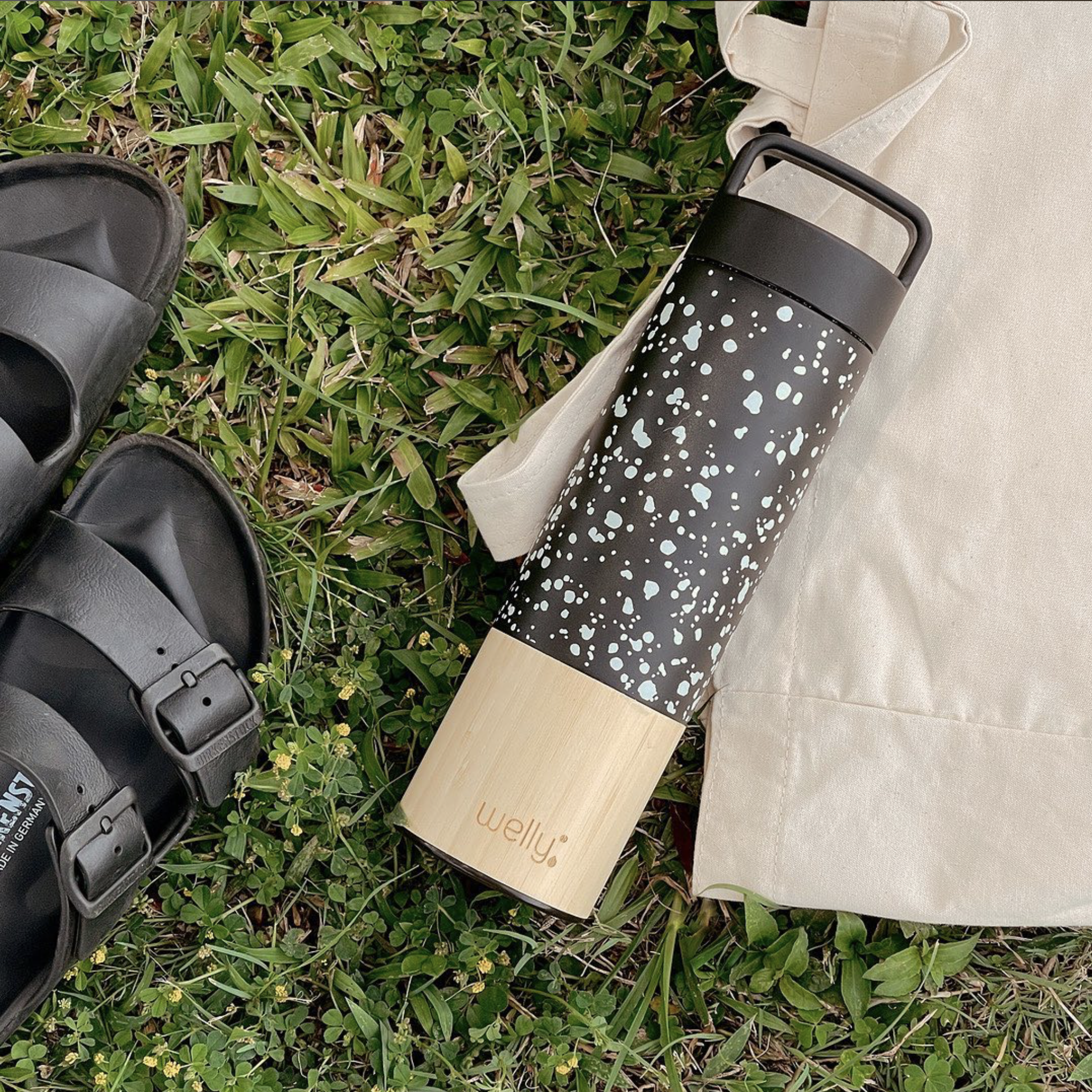 A black and beige Wellys thermos with a splatter paint design lying on grass next to black sandals and a beige cloth bag.