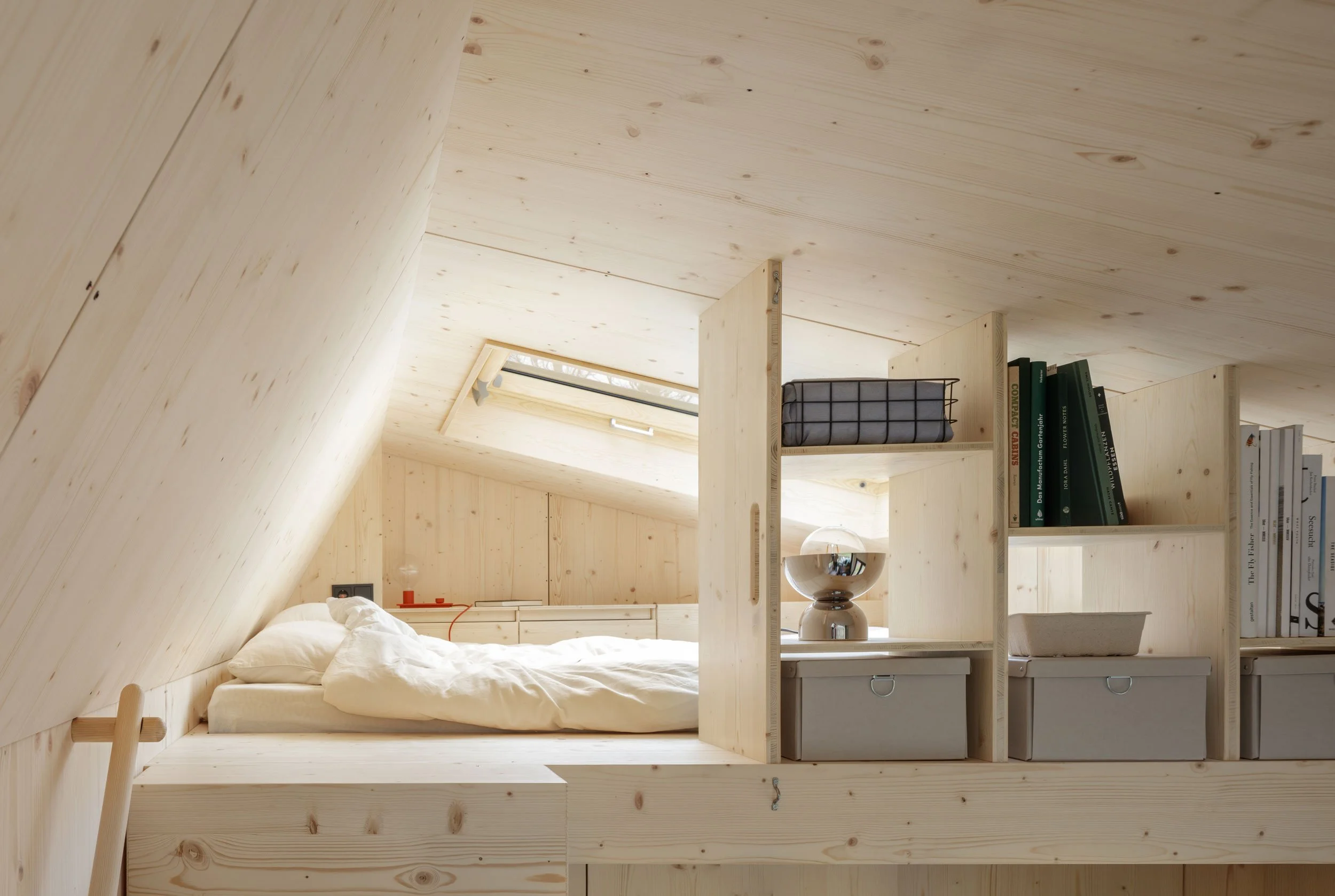 A minimalist bedroom with light wood walls, ceiling, and built-in shelves, featuring a bed, books, storage boxes, a lamp, and a skylight letting in natural light.
