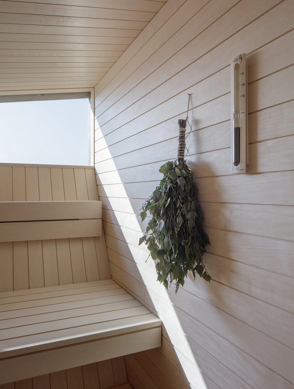 Inside a wooden sauna with a bench, a window allowing natural light, a bundle of leaves hanging on the wall, and a thermometer mounted beside it.