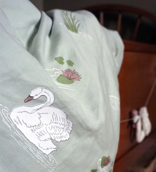 Close-up of light green fabric with a printed handdrawn design of a white swan, water lilies, and lily pads as baby bedding for Gamcha