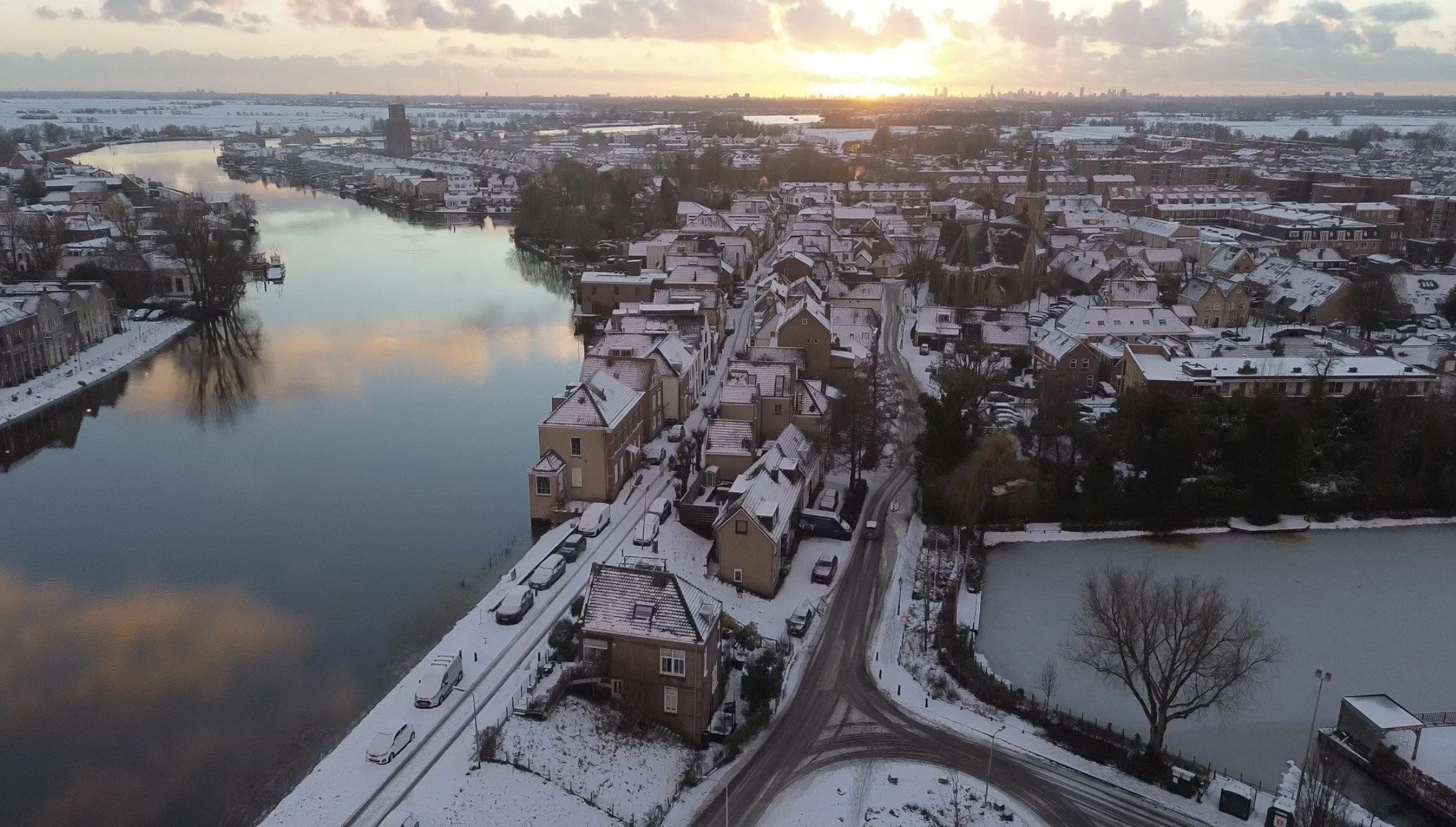Luchtfoto van een winterlandschap met een rivier, een bevroren meer en een woonwijk met sneeuw bedekte huizen, bomen en wegen, bij zonsondergang.