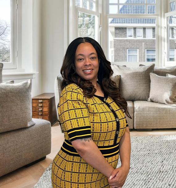 Tammarra Peacock A woman with long dark hair wearing a yellow and black patterned dress, standing in a bright living room with large windows and beige sofas.