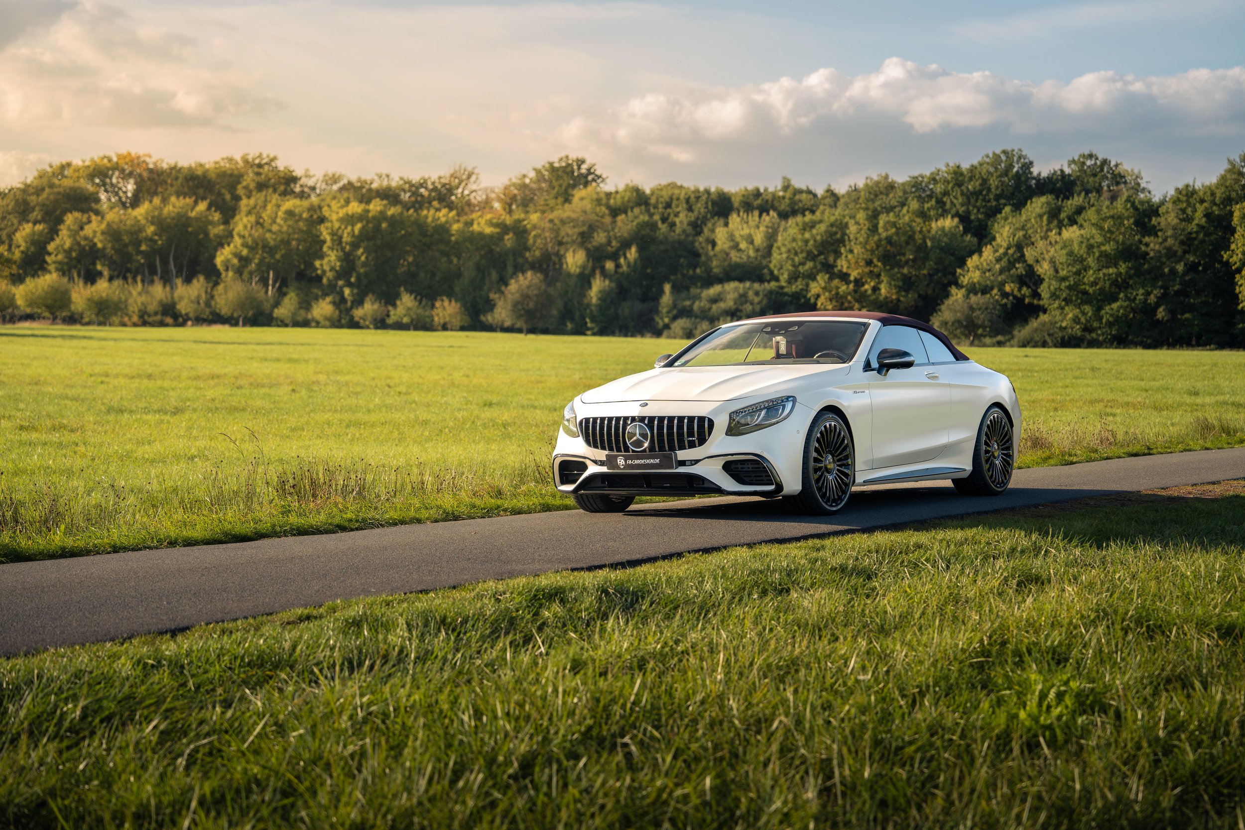Weißer Mercedes-Benz S63 Cabrio mit schwarzen Mansory Felgen auf einer kleinen Straße in einer grünen Landschaft bei Sonnenuntergang.