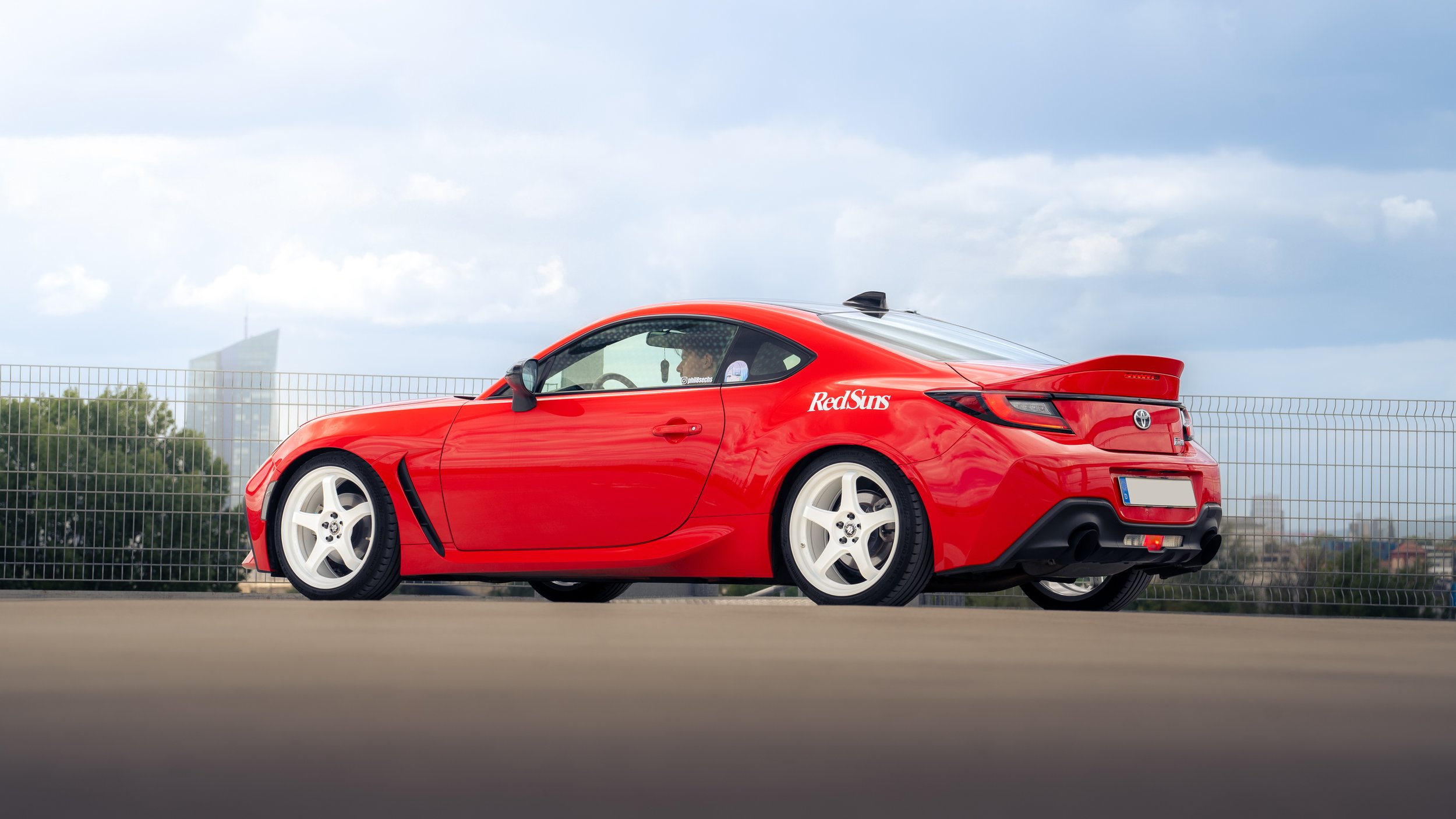 Roter Toyota GR86 mit weißem Felgen, auf einer Parkfläche, mit Stadt im Hintergrund und bewölktem Himmel.