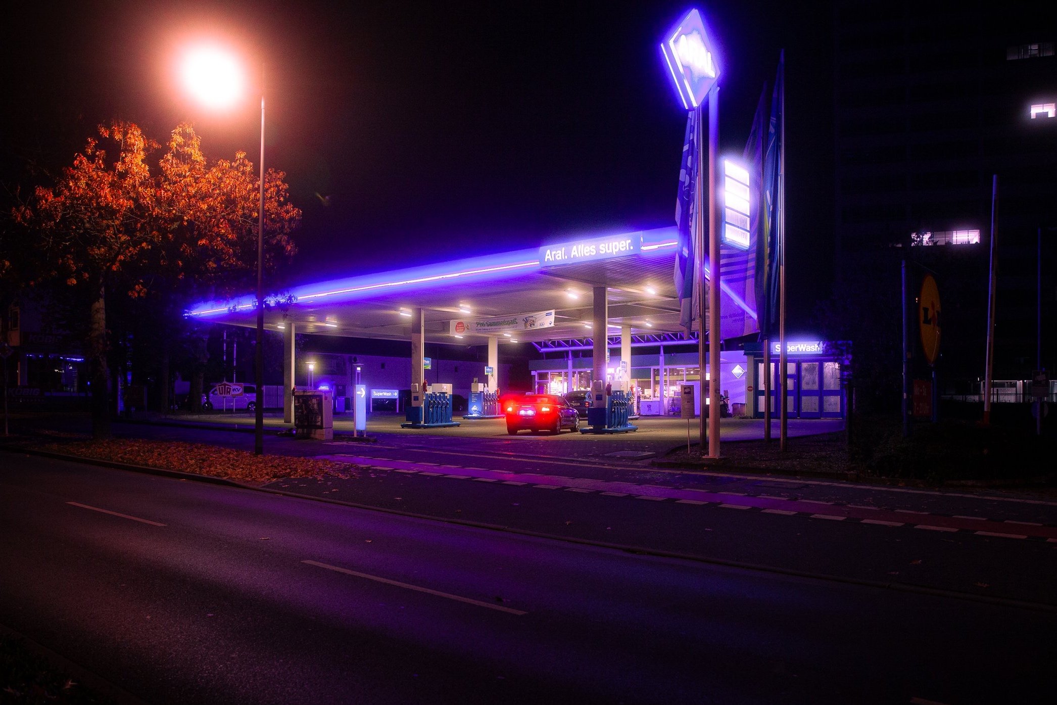 Beleuchtete Tankstelle bei Nacht mit Neonlicht in Blau und Lila, ein Auto tankt, Bäume mit Herbstlaub und Straßenmarkierungen sichtbar.