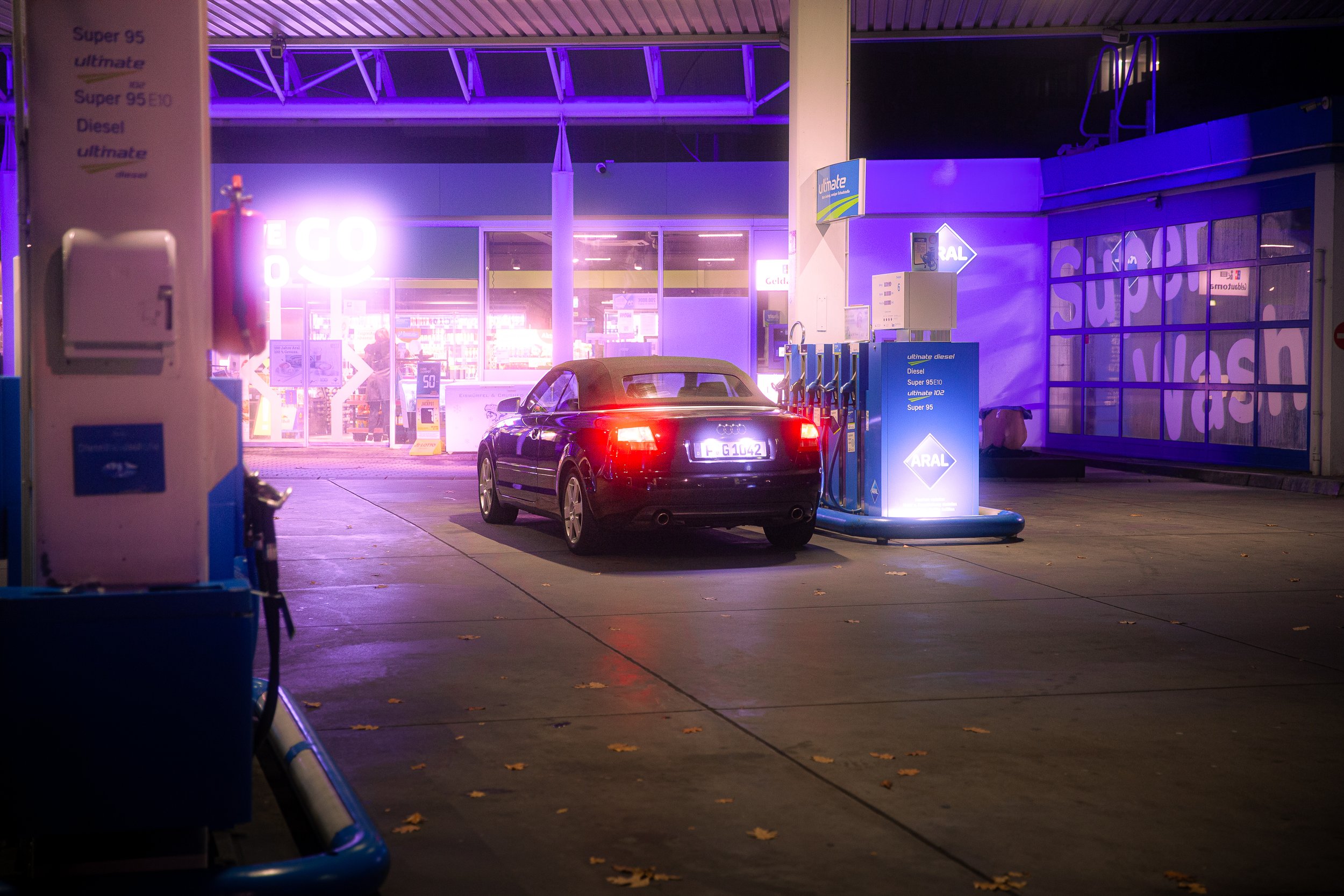 Ein schwarzer Cabriolet steht an der Zapfsäule eines Supermarkttankstellen, im Hintergrund leuchten Neonlichter des Supermarkts bei Nacht, mit einem reflektierenden blauen und violetten Licht.