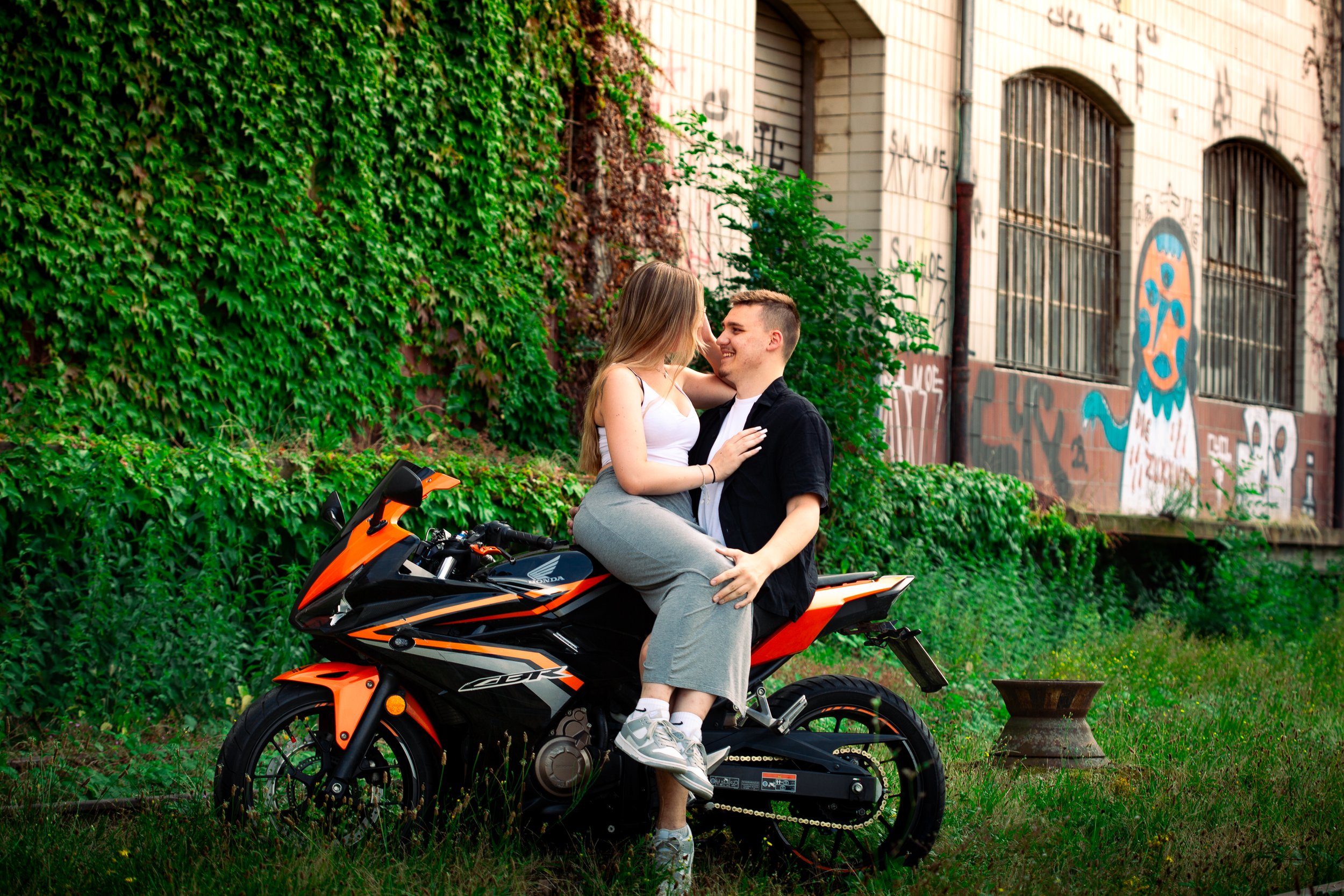 Ein junges Paar sitzt auf einem schwarzen und orangefarbenen Motorrad vor einer Wand mit Graffiti, umgeben von grünen Pflanzen, die sich an der Wand hochziehen, und schaut sich verliebt an.