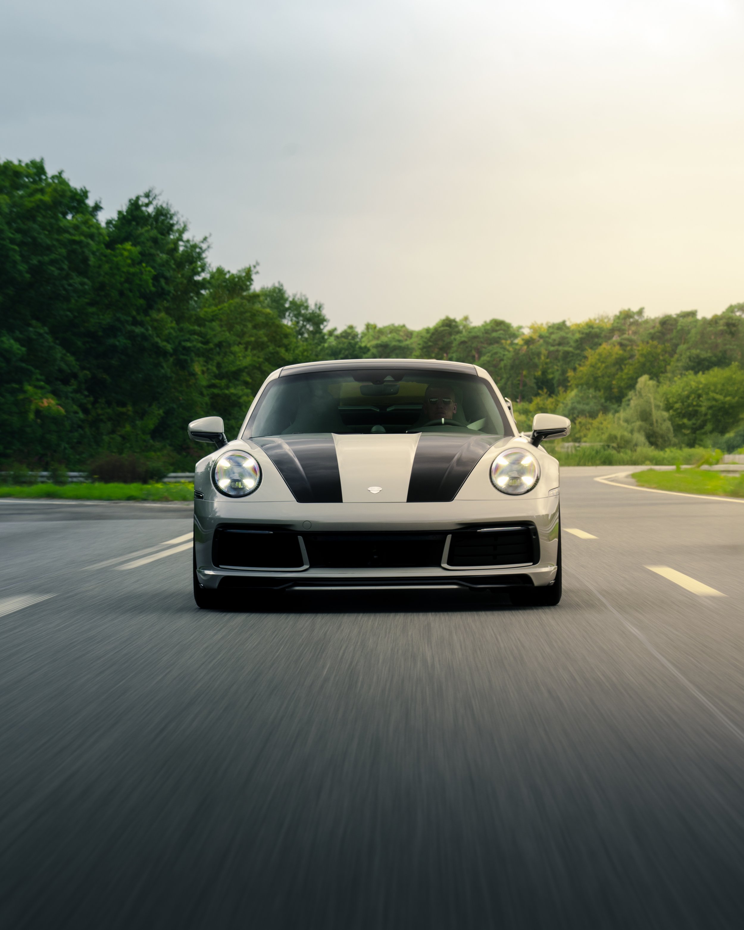 Ein kreidefarbener Porsche 992 Carrera S Techart fährt auf einer Autobahn, umgeben von grünen Bäumen und einem Himmel mit Wolken.