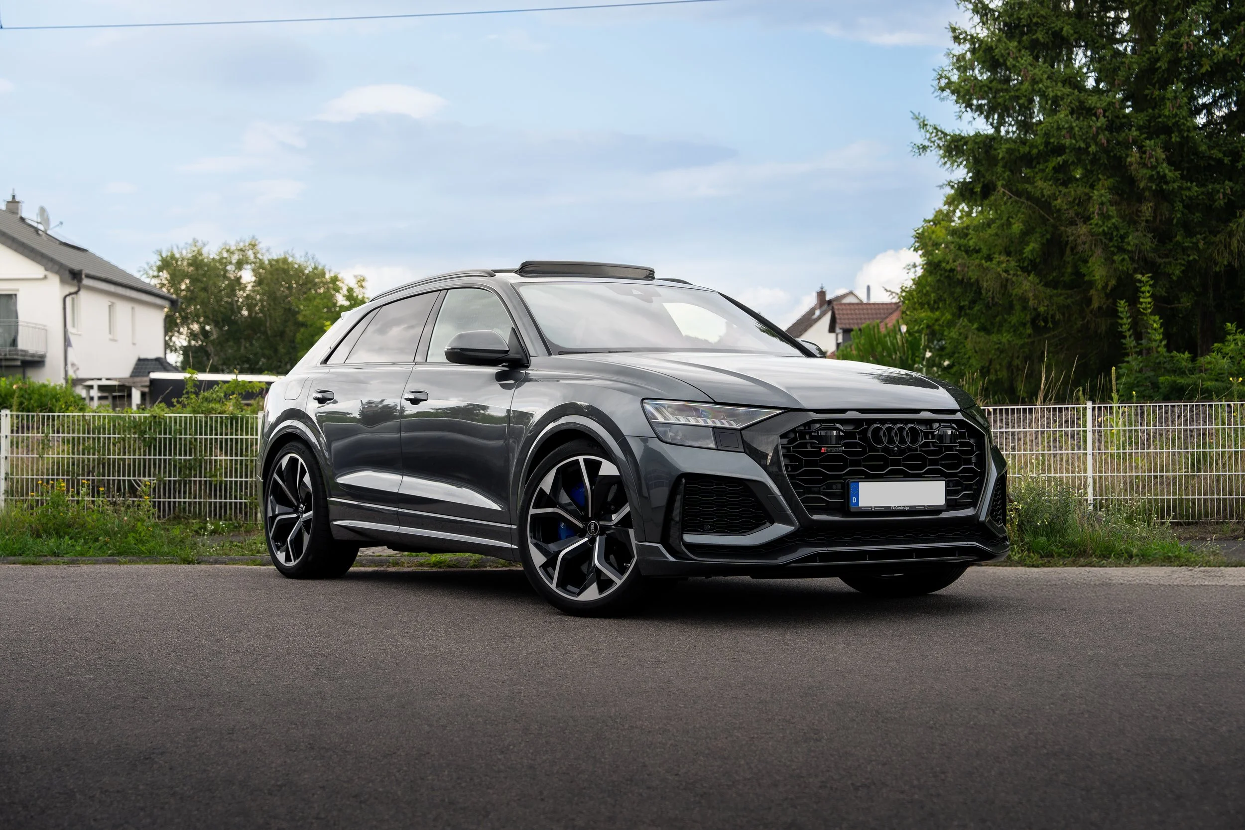 Ein grauer Audi RS Q8 auf einer asphaltierten Straße vor einem Zaun, im Hintergrund Häuser und Bäume.