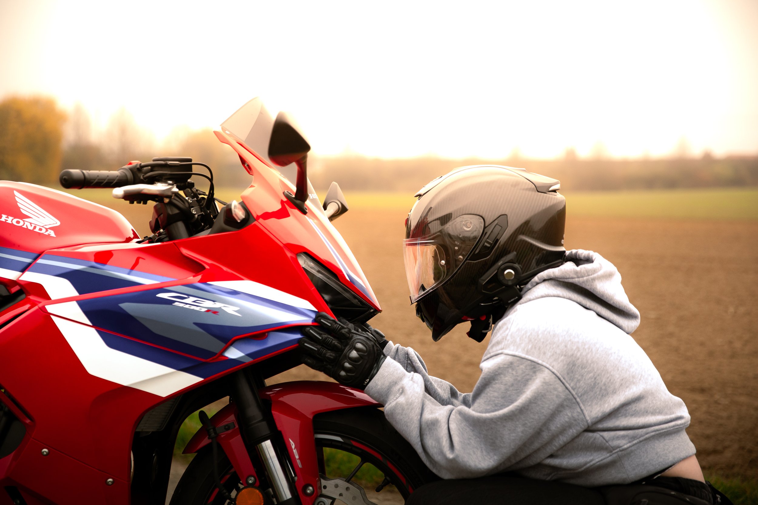 Person in Motorradhelm und grauem Pullover betrachtet ein rotes Honda Motorrad im Freien auf einem landwirtschaftlichen Feld, bei Sonnenuntergang.