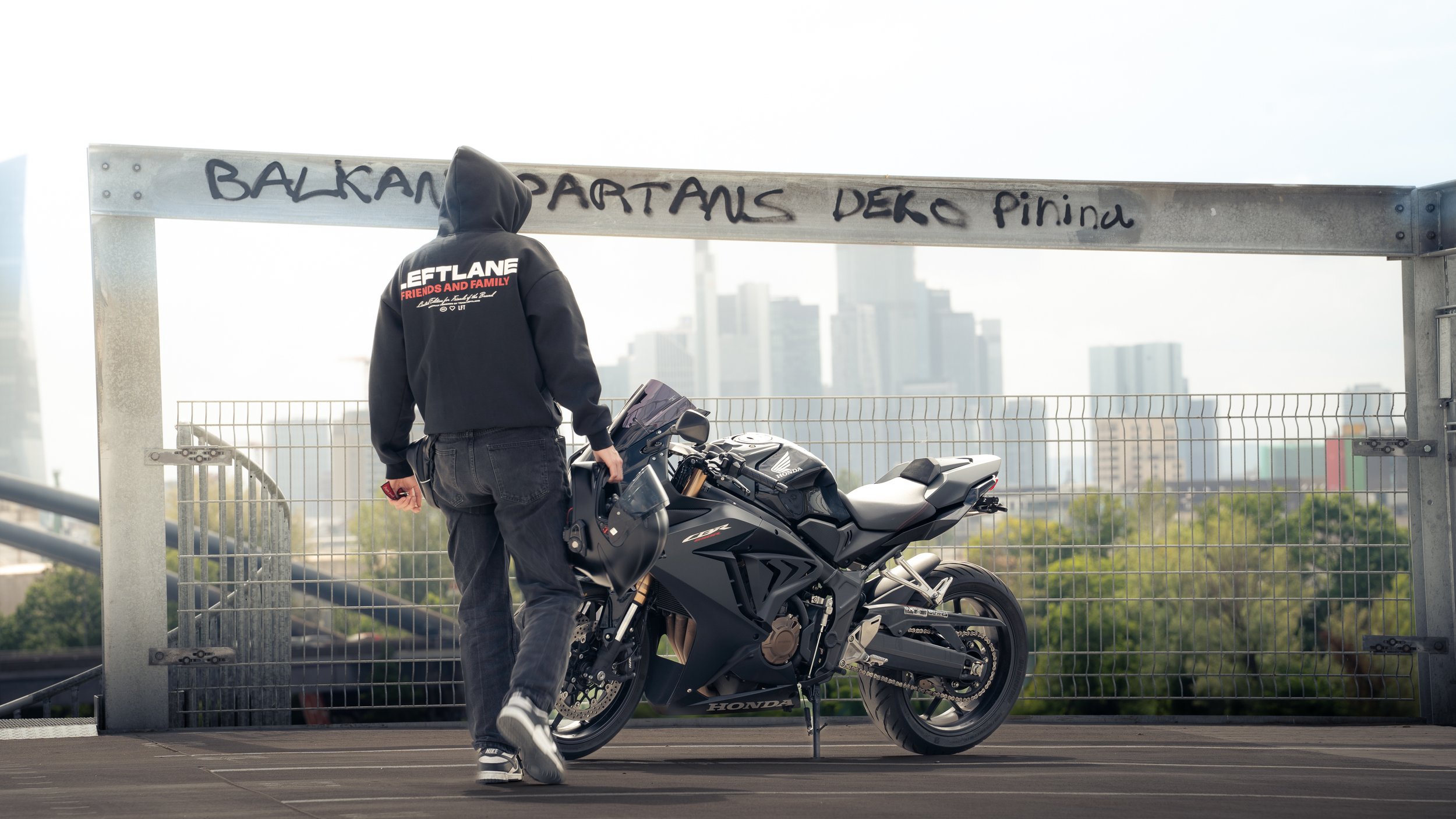 Eine Person mit Kapuzenjacke, die auf einem Parkdeck neben einer schwarzen Honda CBR 650 R steht. Im Hintergrund eine städtische Skyline mit Hochhäusern.