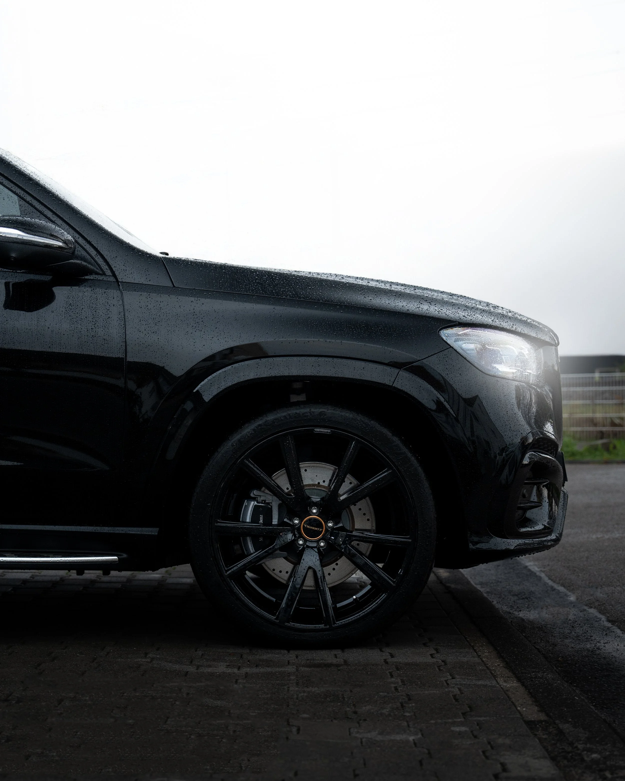 Nahaufnahme eines schwarzen Mercedes GLS 450d mit schwarzen Mansory Felgen, der bei Regen auf einem gepflasterten Parkplatz steht, zeigt das vordere rechte Rad und einen Teil der Karosserie.