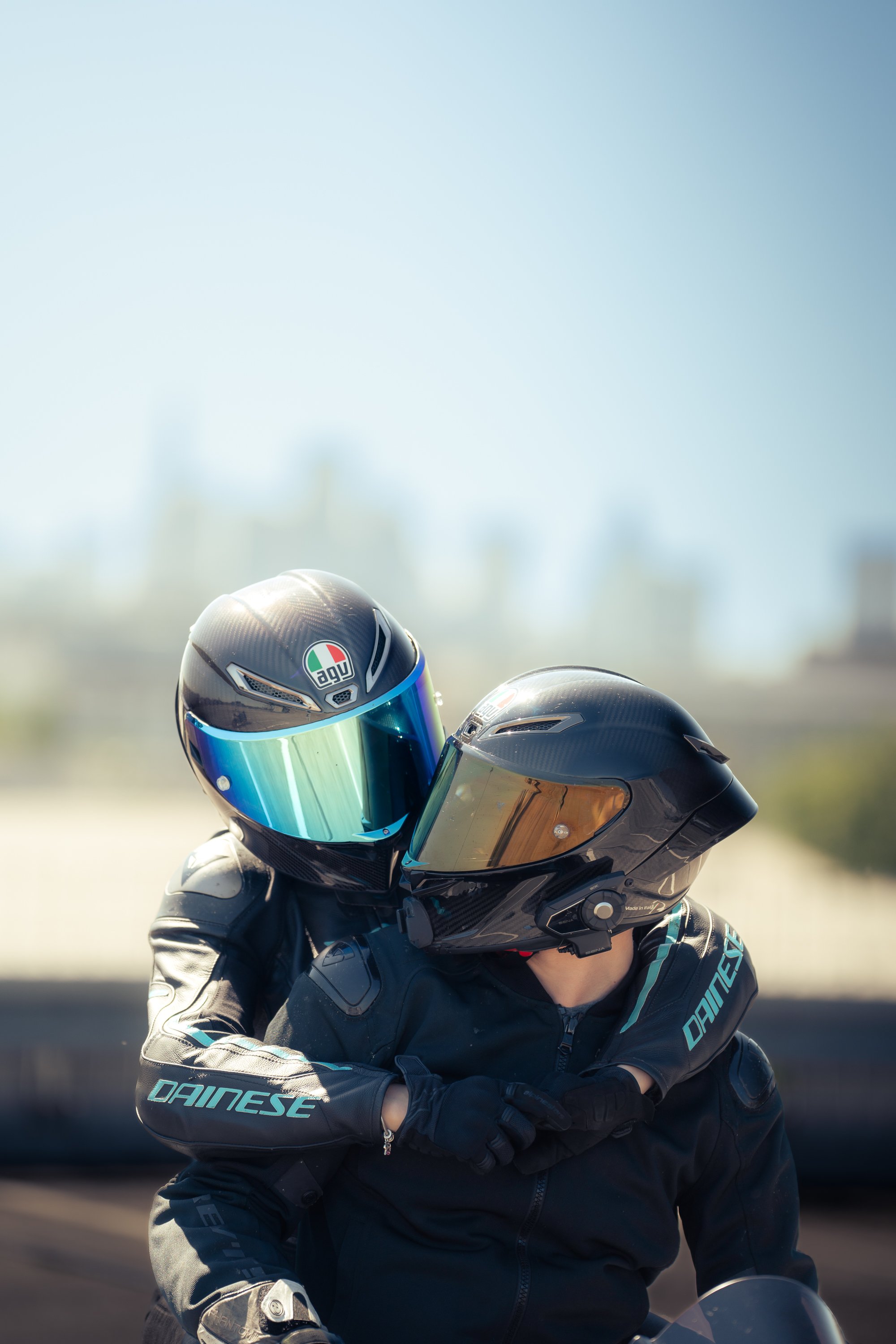 Zwei Motorradfahrer in Schutzhelmen und Motorradausrüstung, einer sitzt auf einem Motorrad, während der andere steht und sich anlehnt.
