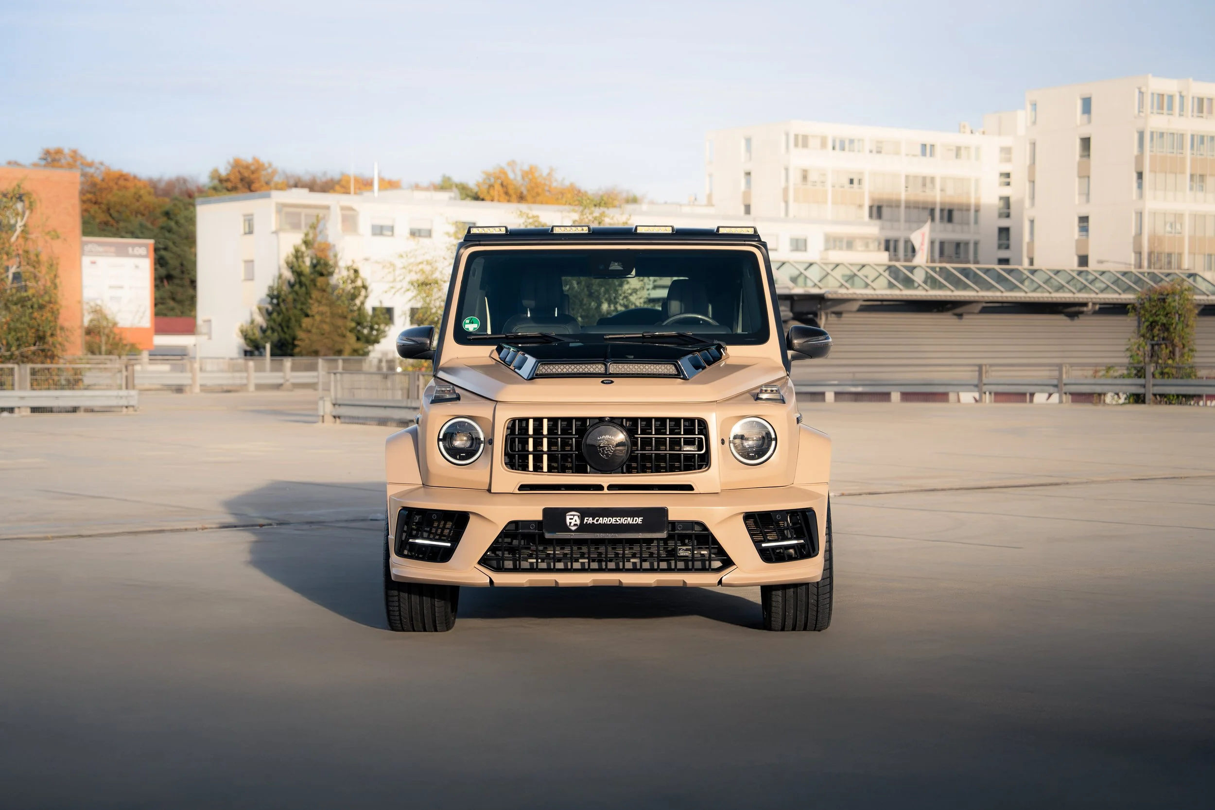 Ein beigefarbener Mercedes G500 mit komplettem Lumma Umbau steht in einem leeren Parkhaus mit modernen Gebäuden im Hintergrund.