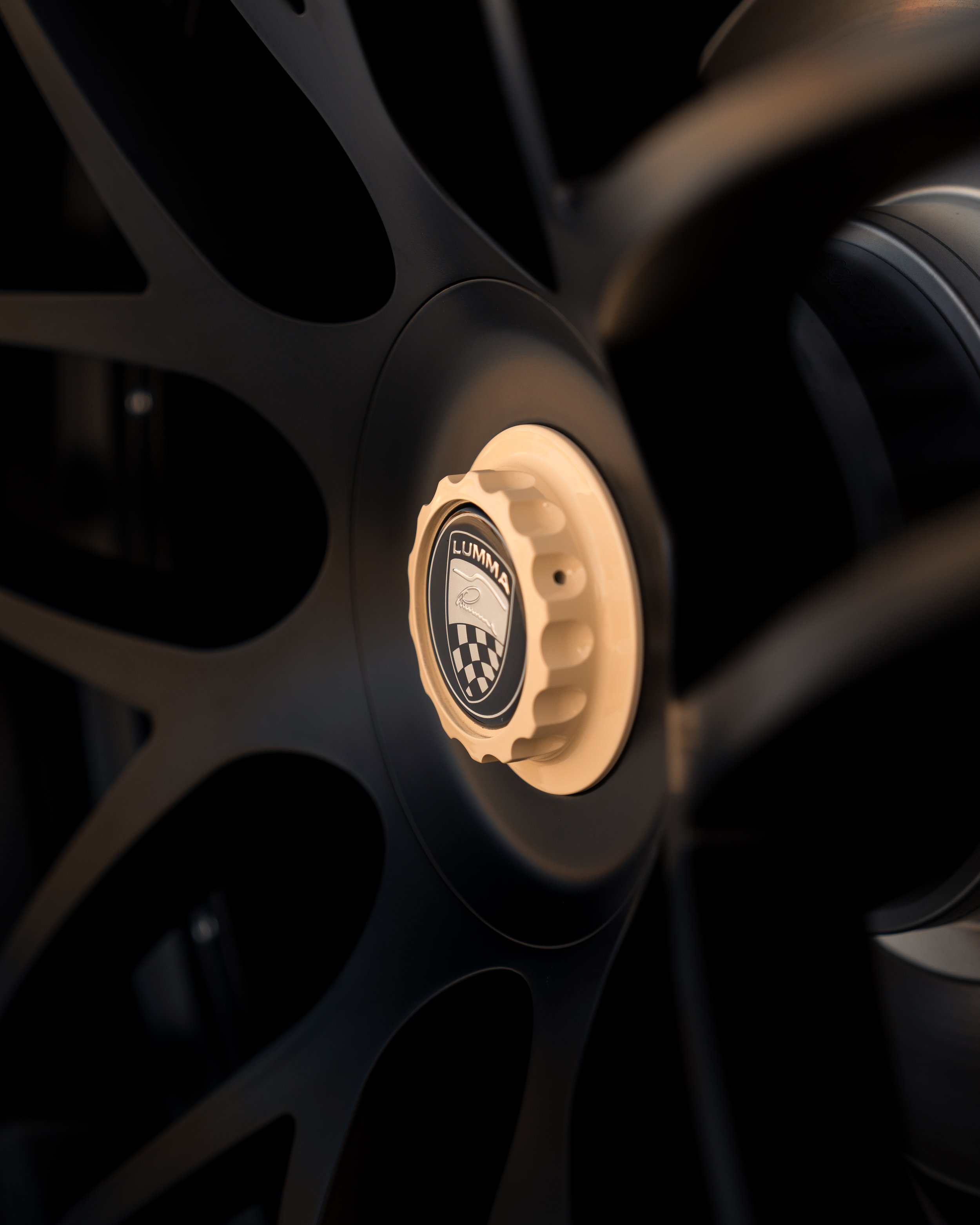 Nahaufnahme einer schwarzen Lumma Felge mit einem zentralen beigefarbenen Nabendeckel mit dem Logo "LUMMA" in Silber und einem schwarzen und weißen Wappen darunter.
