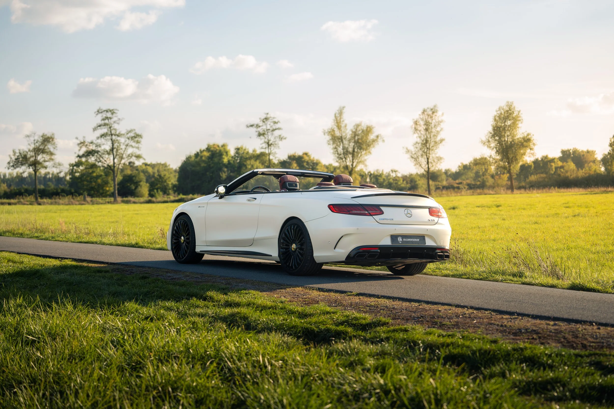 Weißer Mercedes-Benz S63 Cabrio mit schwarzen Mansory Felgen auf einer Landstraße bei Sonnenuntergang, umgeben von grünen Wiesen und Bäumen.