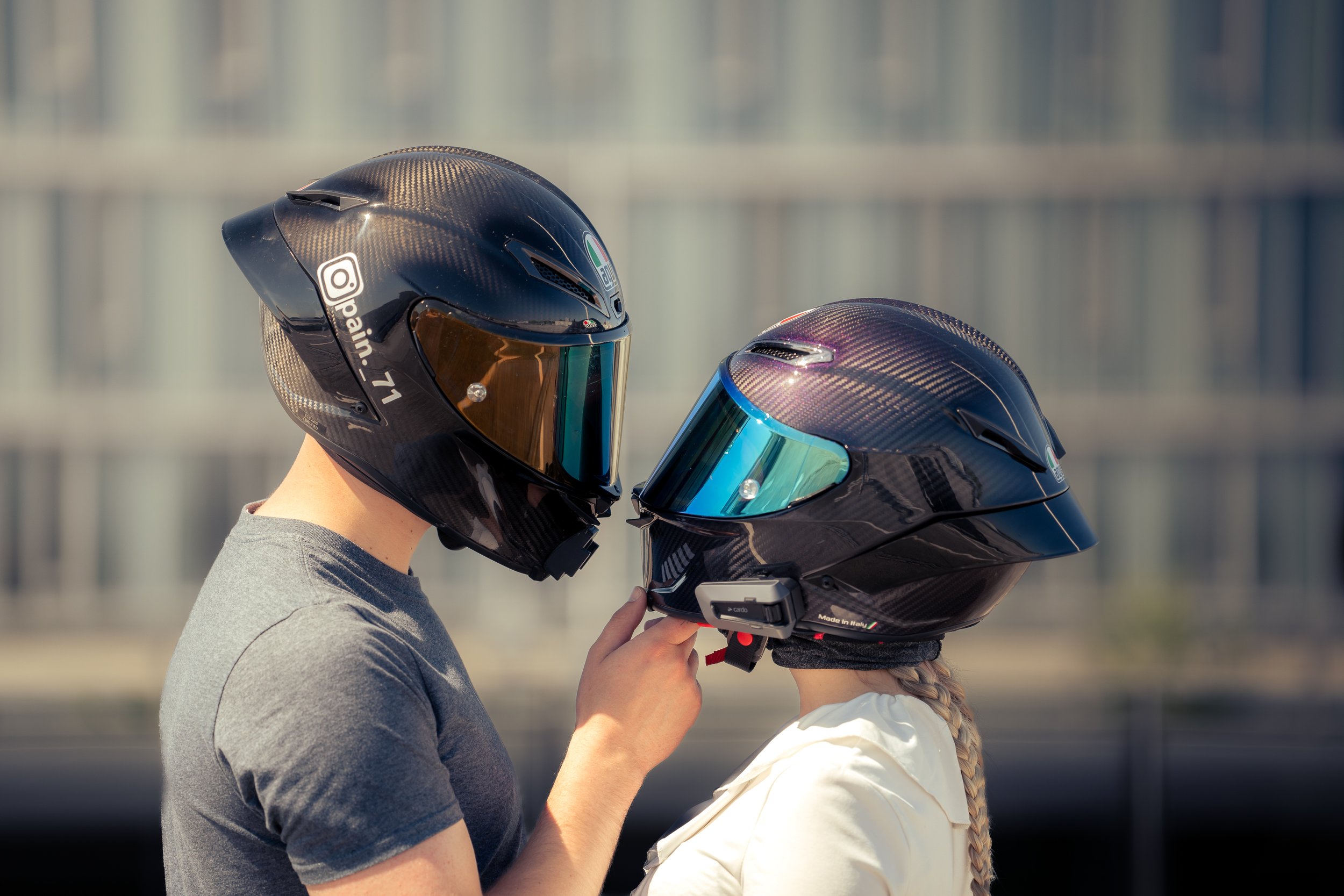 Zwei Personen tragen schwarze Motorradhelme und stehen sich gegenüber, während sie ihren Helme anpassen, auf einer Straße mit urbanem Hintergrund.